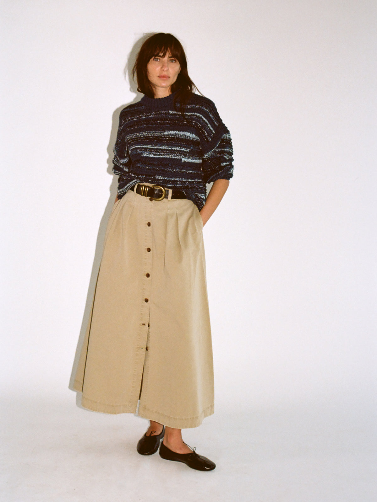 A woman stands against a plain white studio background wearing a dark navy and gray striped knit sweater, a long beige button-front skirt, a brown belt, and dark loafers., image 6 of 6