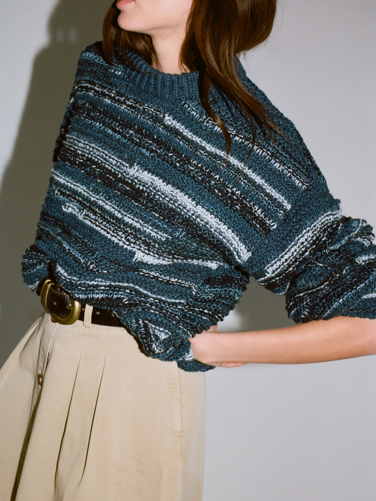 A woman wearing a chunky knit sweater with horizontal stripes in teal, black, and white, paired with beige trousers and a brown belt., image 2 of 6