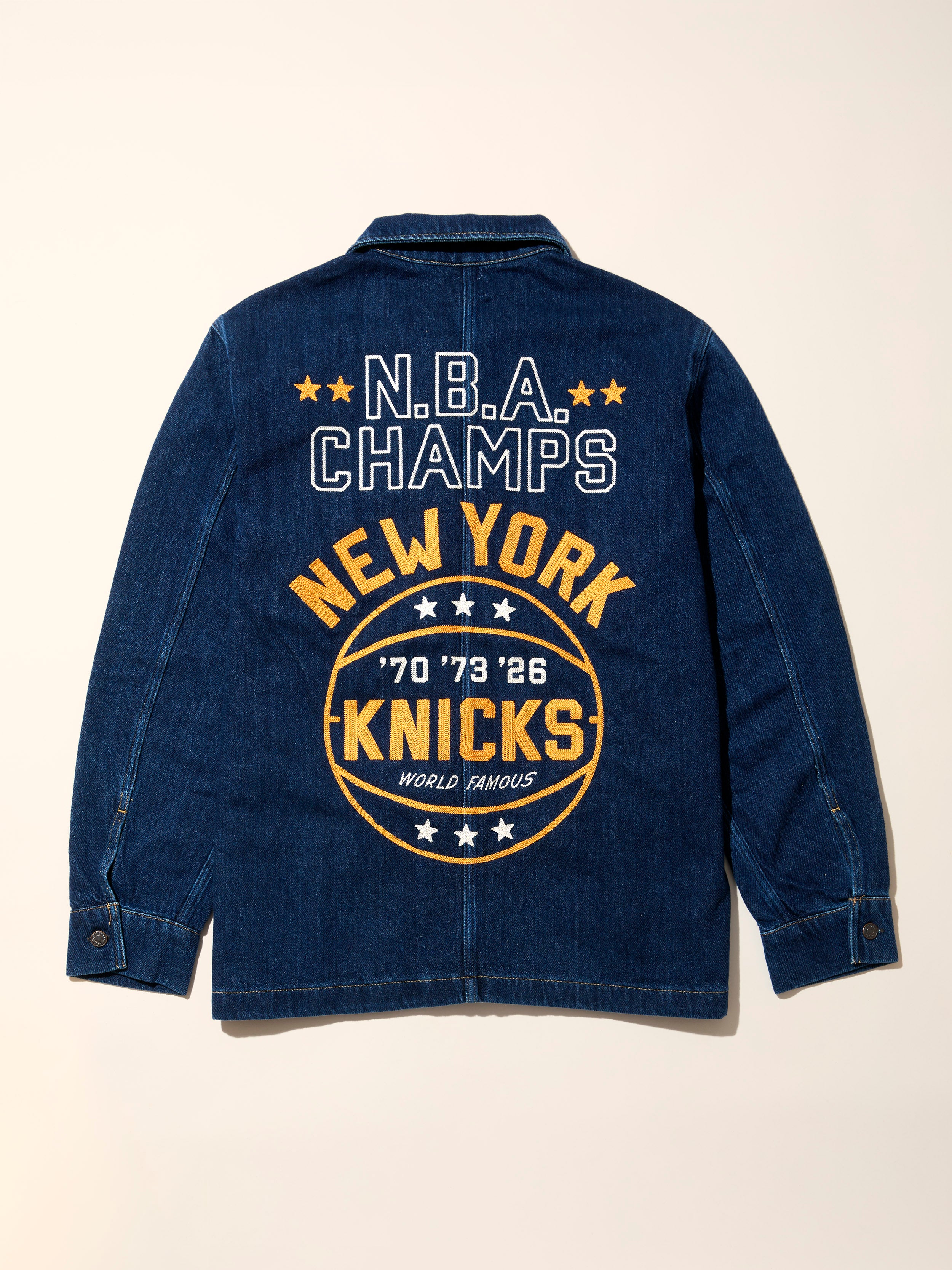 Blue denim jacket with embroidered N.B.A. Champs New York Knicks graphic on the back against a beige background