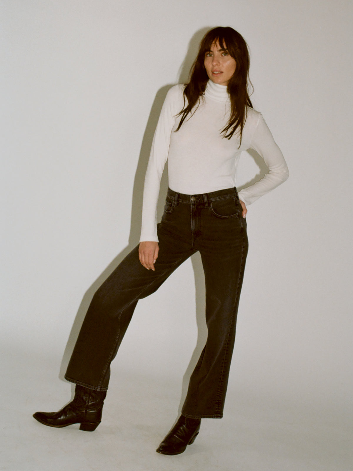A model wearing a white turtleneck knit with black denim jeans and black boots standing against a plain light gray studio background., image 7 of 7