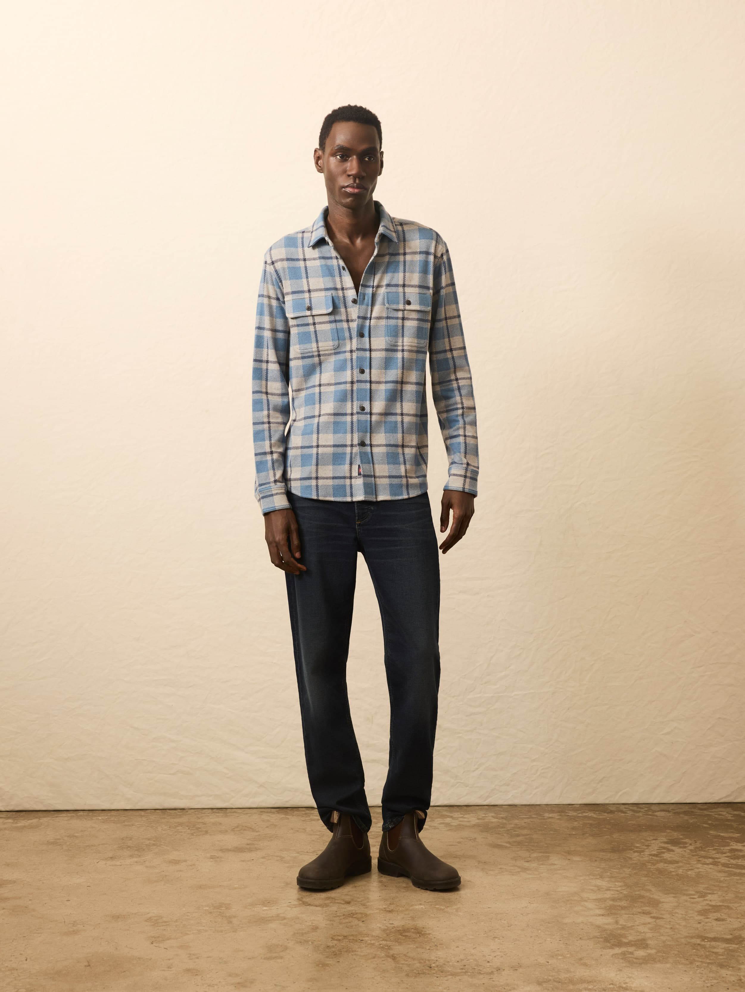 Man wearing a blue plaid shirt and dark jeans standing against a beige wall.