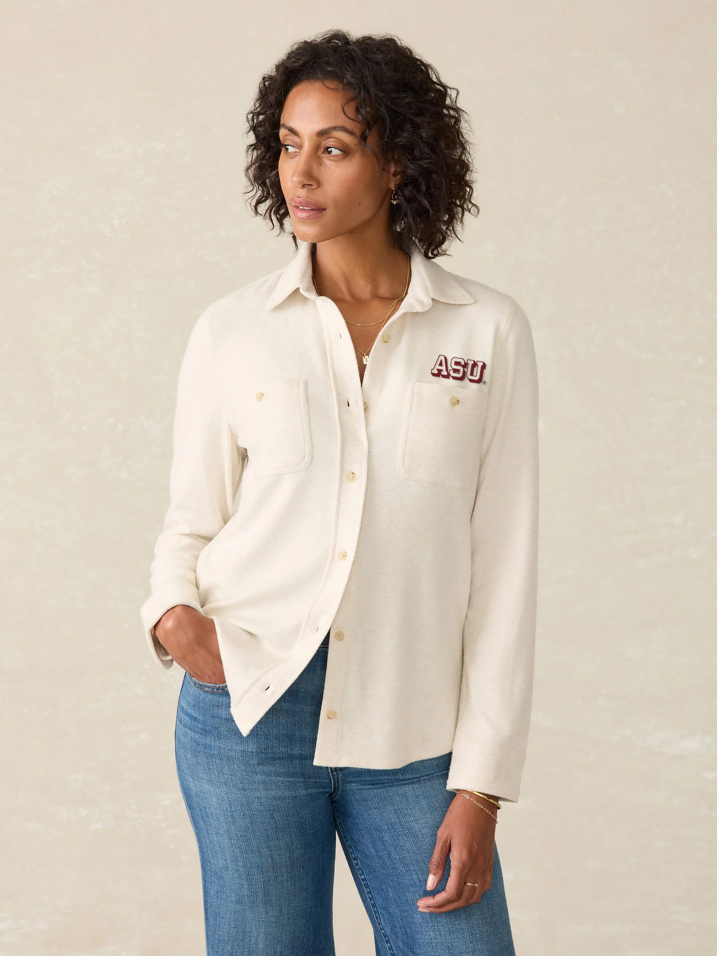 Woman wearing a cream-colored button-up shirt with Arizona State University logo on the left chest and blue jeans on a beige background