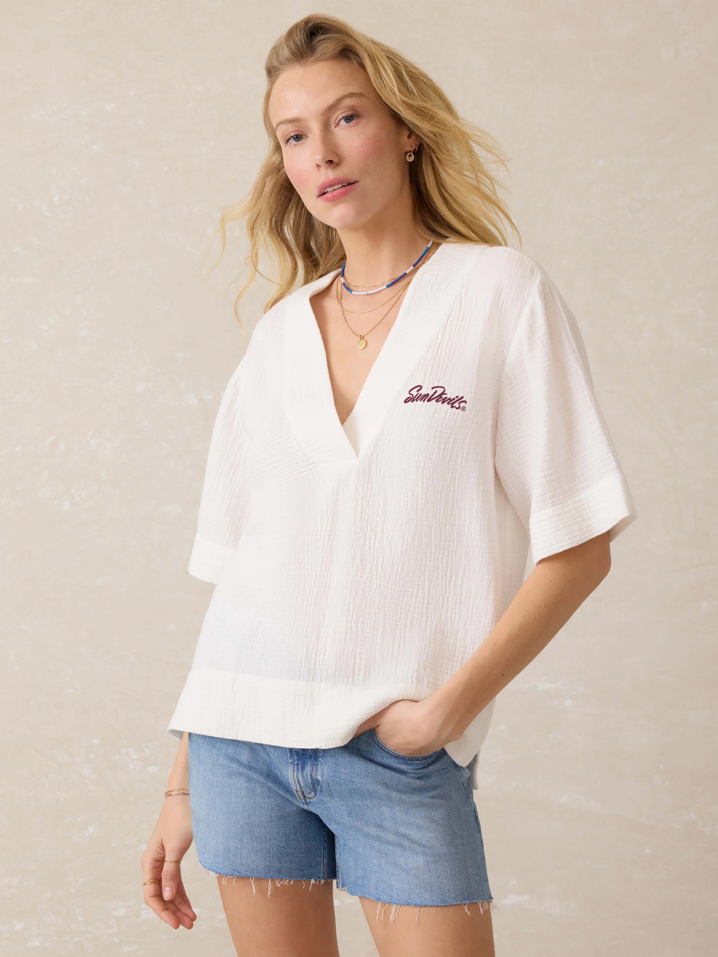 Woman wearing a white blouse with an Arizona State University graphic on the left chest and denim shorts against a beige background
