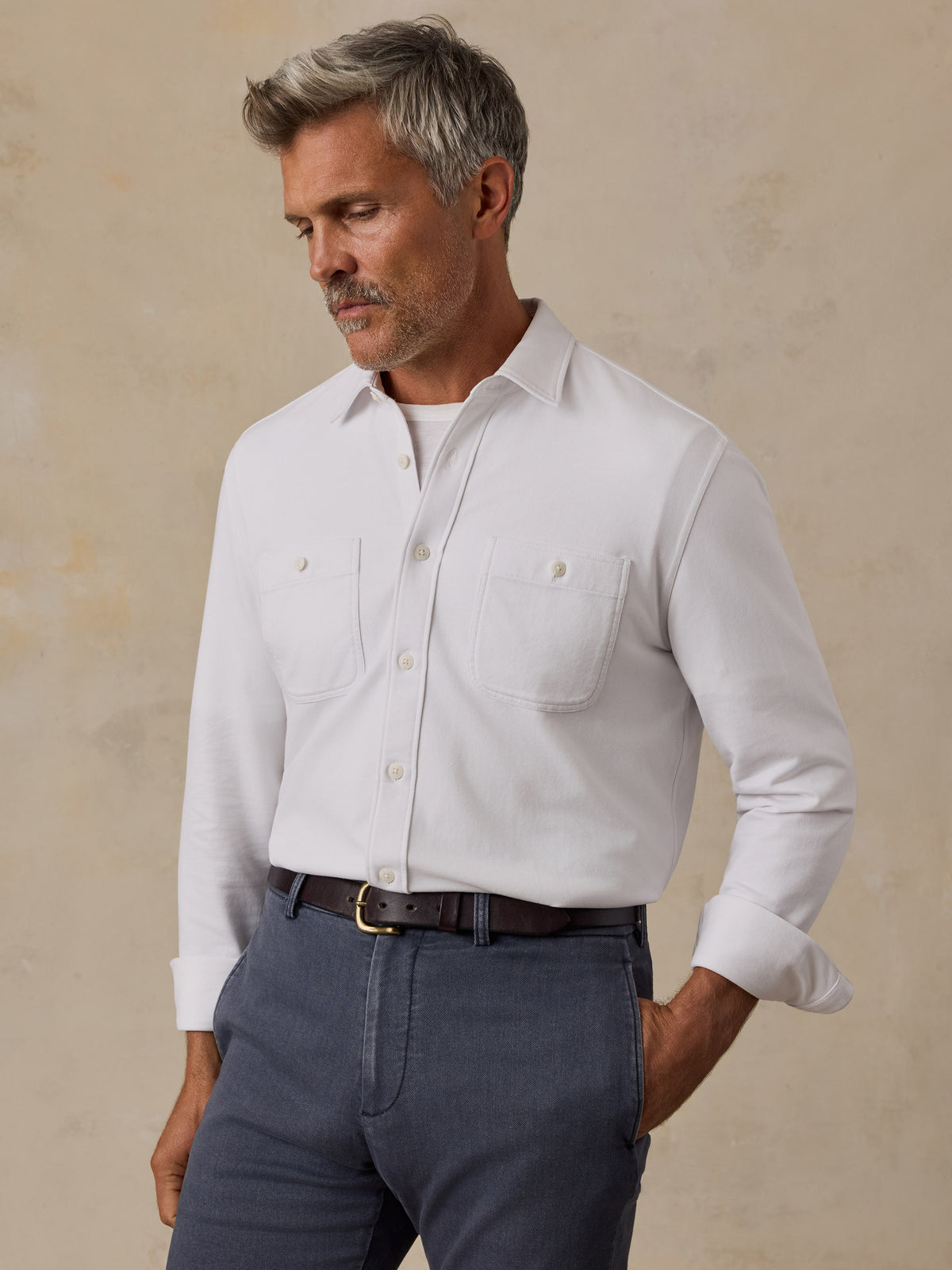 A man wearing a white long-sleeve button-down shirt with two chest pockets, tucked into dark gray trousers with a brown belt, standing against a textured beige background., image 1 of 6