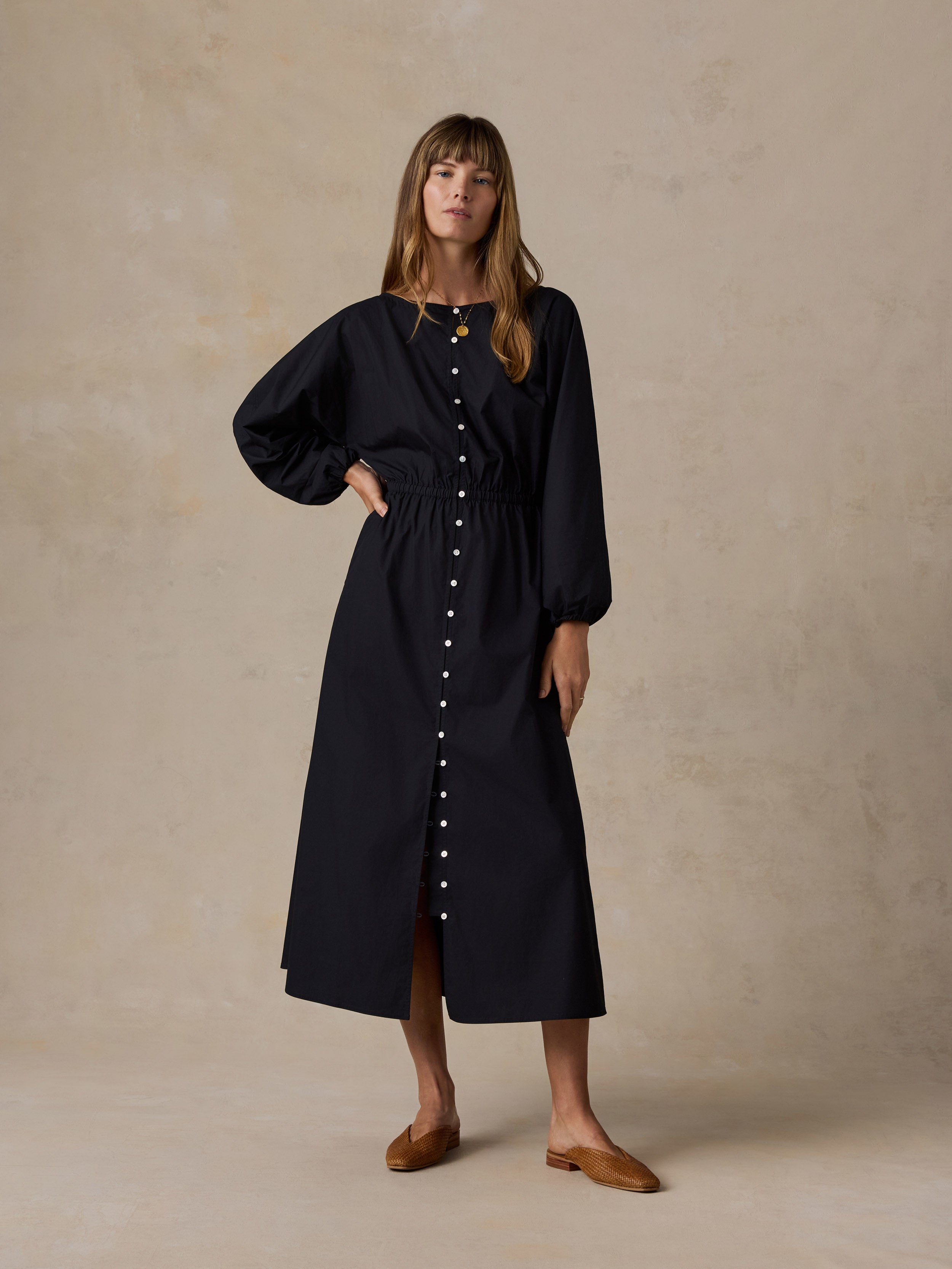 A woman modeling a long black midi dress with a button-down front, gathered waist, and voluminous sleeves, standing against a textured beige studio backdrop.