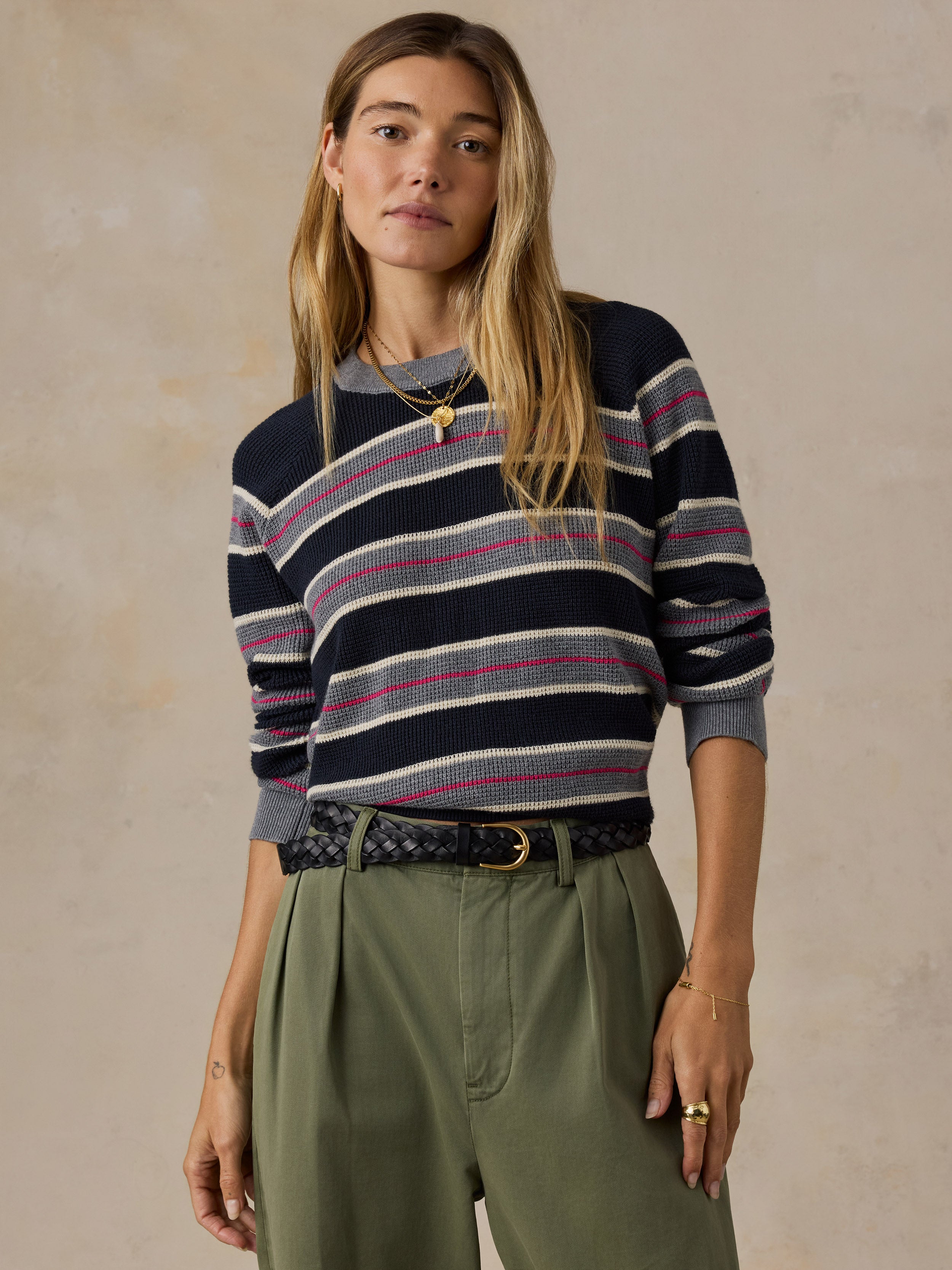 A woman modeling a navy blue crewneck sweater with horizontal gray, white, and pink stripes, paired with olive green trousers and a patterned belt, set against a textured beige studio background.