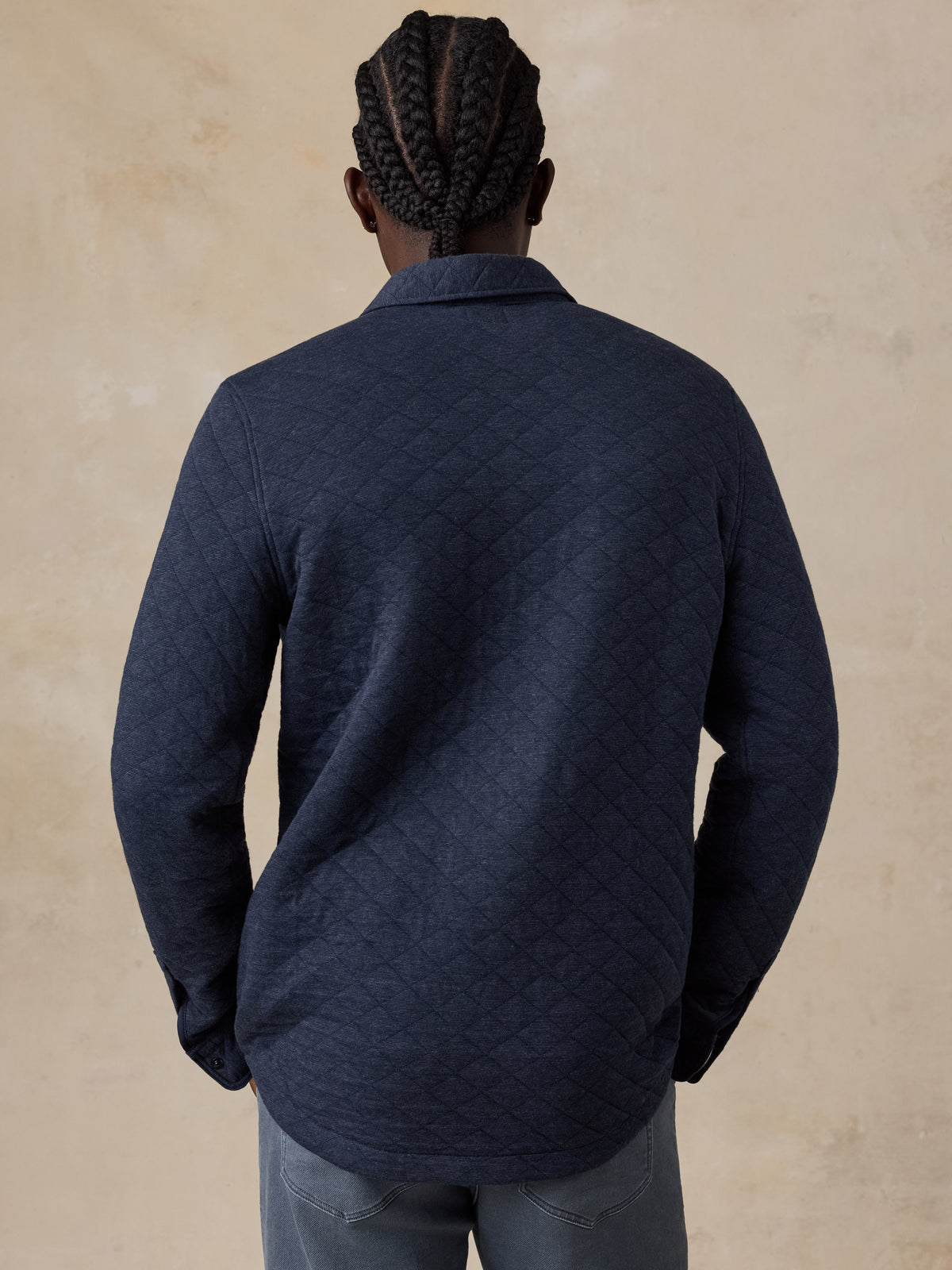 A man viewed from behind wearing a dark navy quilted long-sleeve jacket and gray jeans, standing against a textured beige background., image 4 of 7