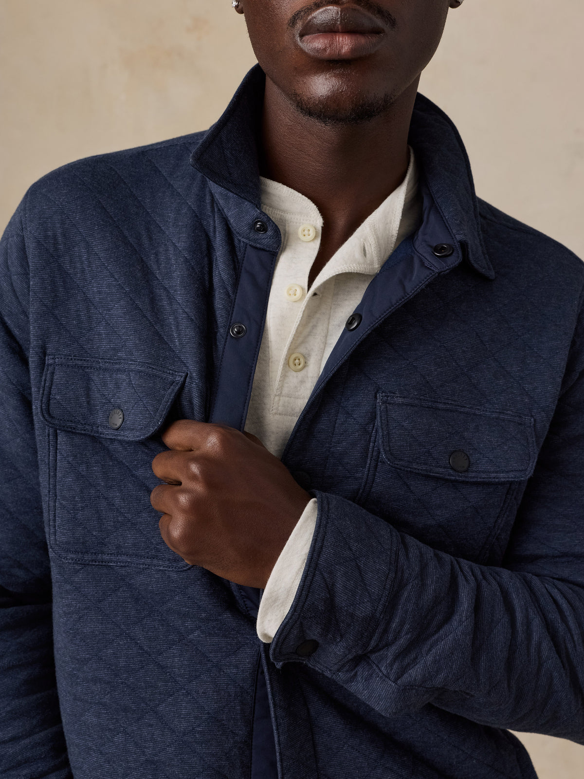 A close-up of a man wearing a textured navy blue overshirt over a cream-colored collared shirt, set against a warm beige background., image 3 of 7