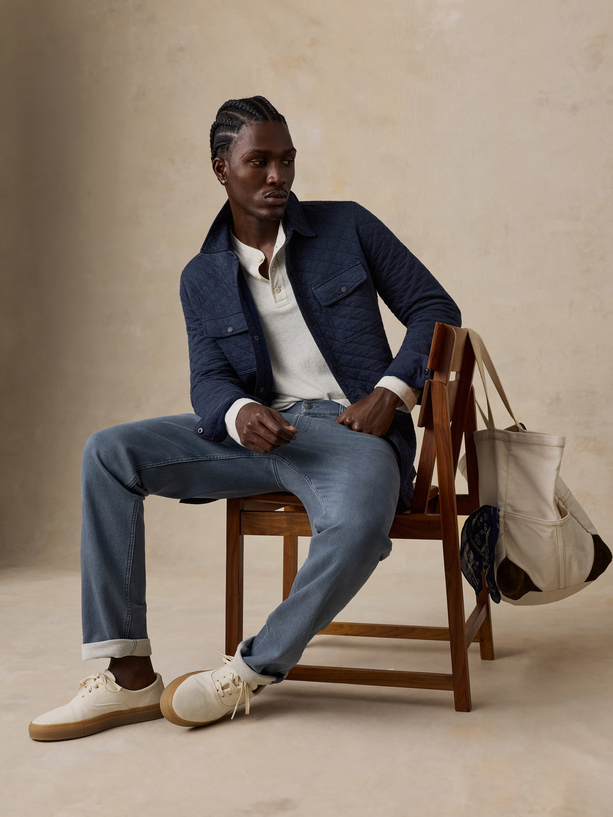A man sits on a wooden chair wearing a navy quilted jacket, light blue jeans, and white sneakers, with a cream canvas tote bag resting on the chair against a warm beige studio backdrop., image 5 of 7