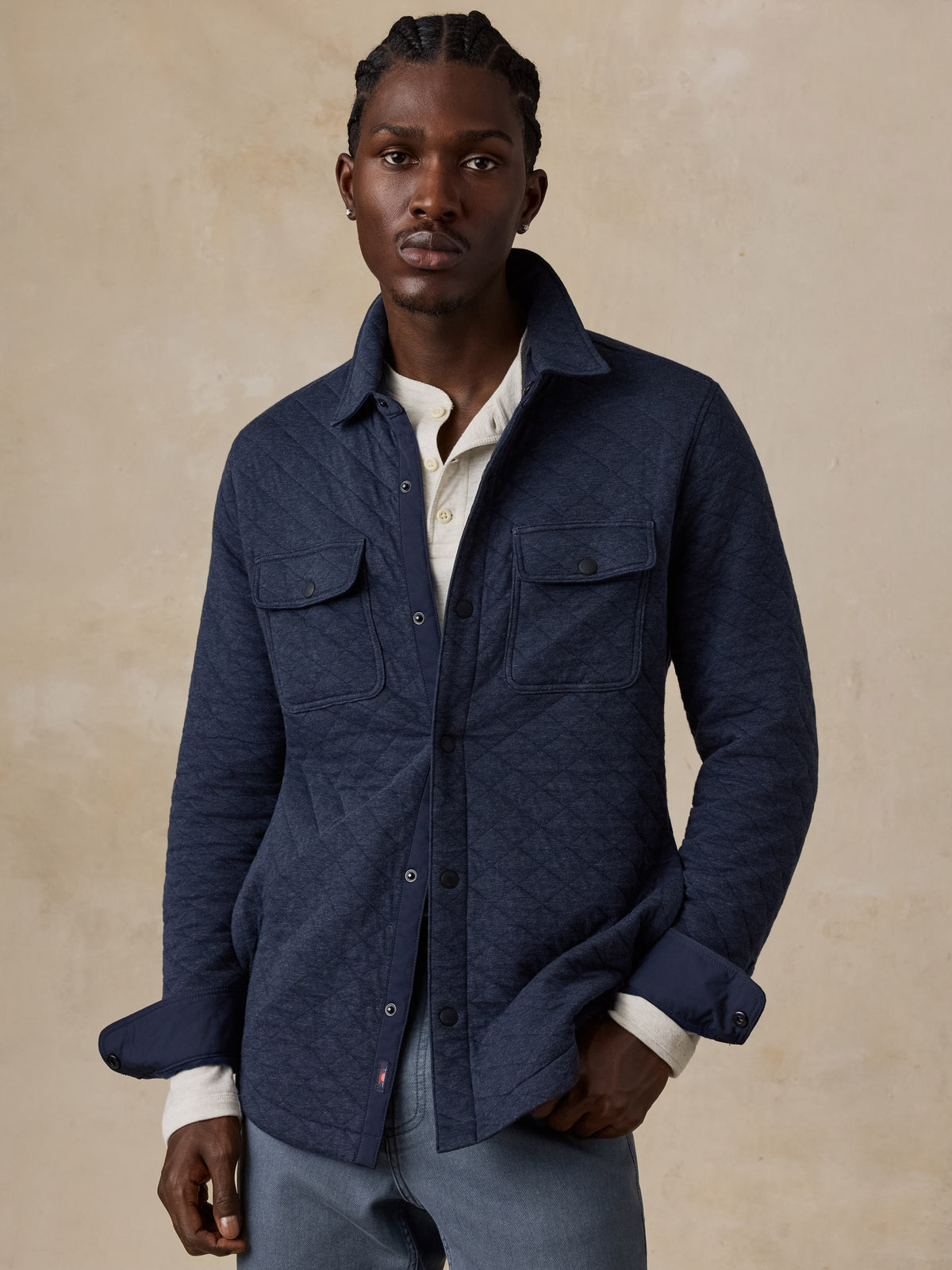 A man wearing a dark navy blue textured overshirt jacket over a white collared shirt and gray trousers, standing against a warm beige textured studio background., image 1 of 7
