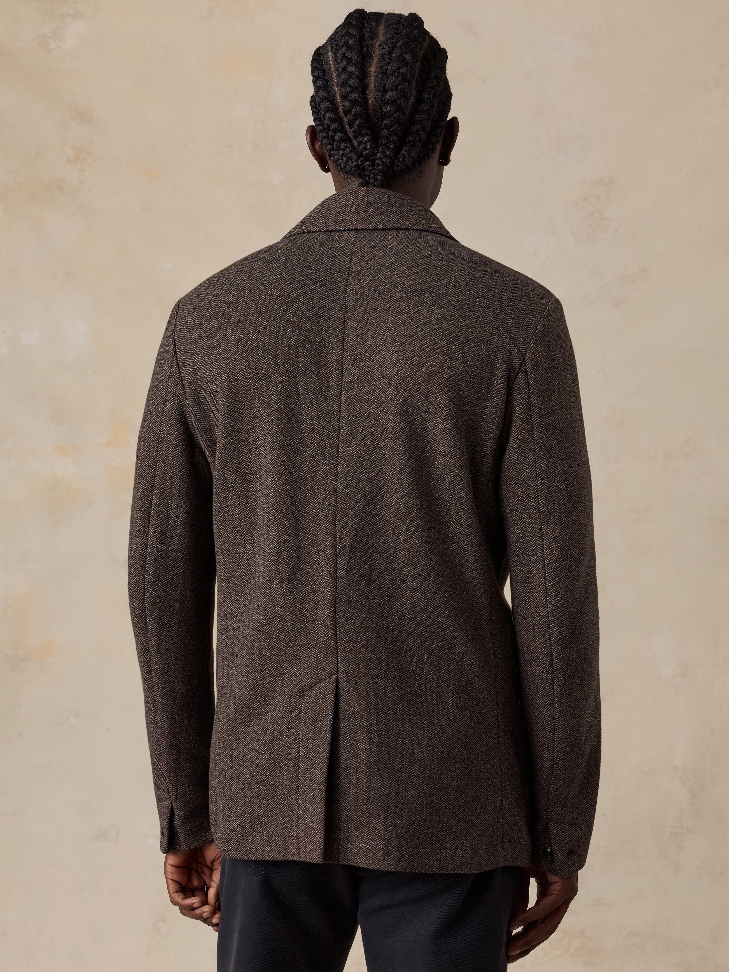 A man viewed from behind wearing a dark brown textured tweed blazer and dark trousers, standing against a textured beige background.
