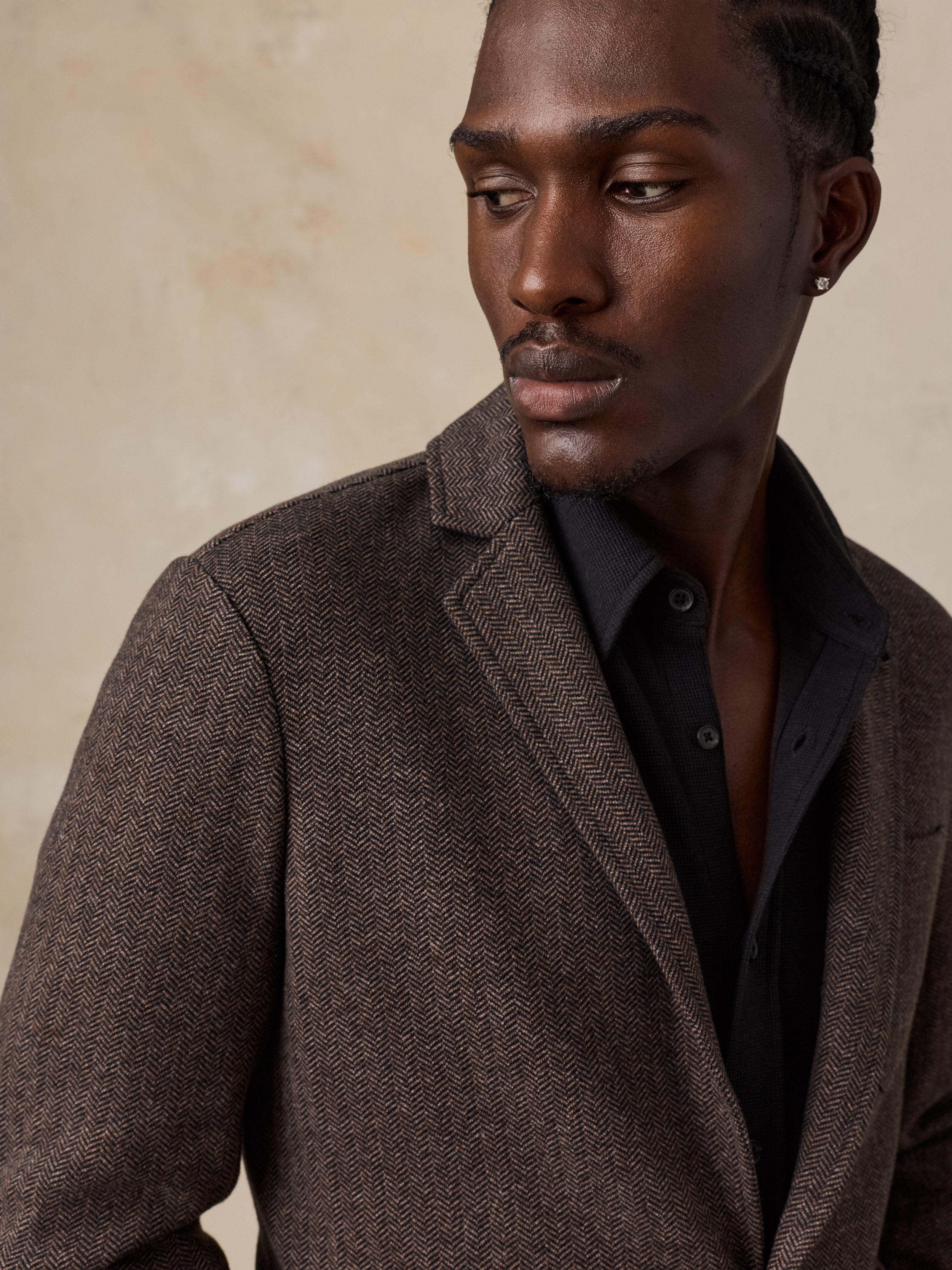 A close-up of a man wearing a brown herringbone blazer over a light blue button-down shirt, with his hands adjusting the blazer's front button.
