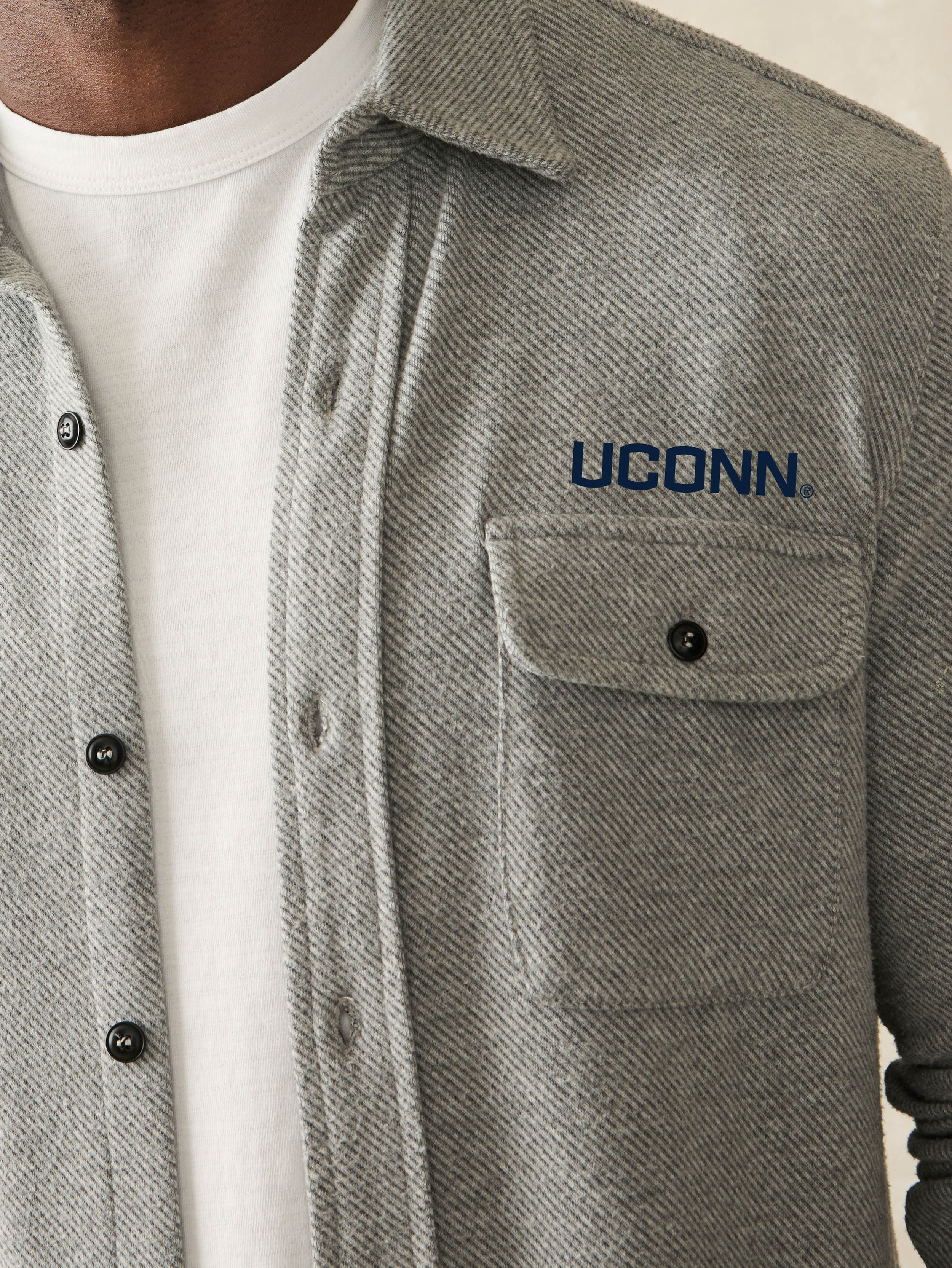 Man is wearing a grey long sleeve shirt with UCONN graphic on left chest and left arm against a beige background