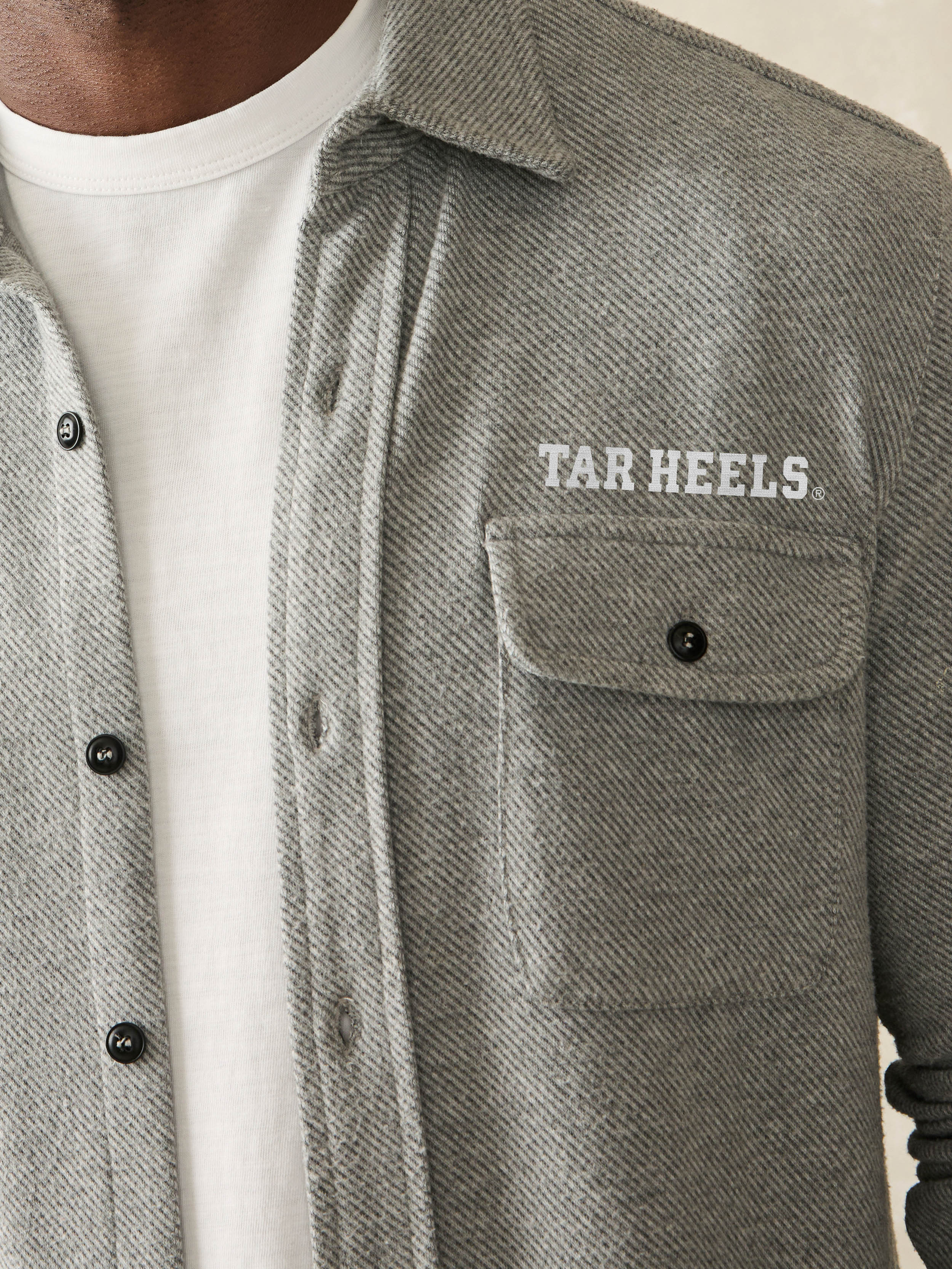 Man wearing a grey long sleeve button up shirt with UNC graphic at the left chest and left arm against a beige background