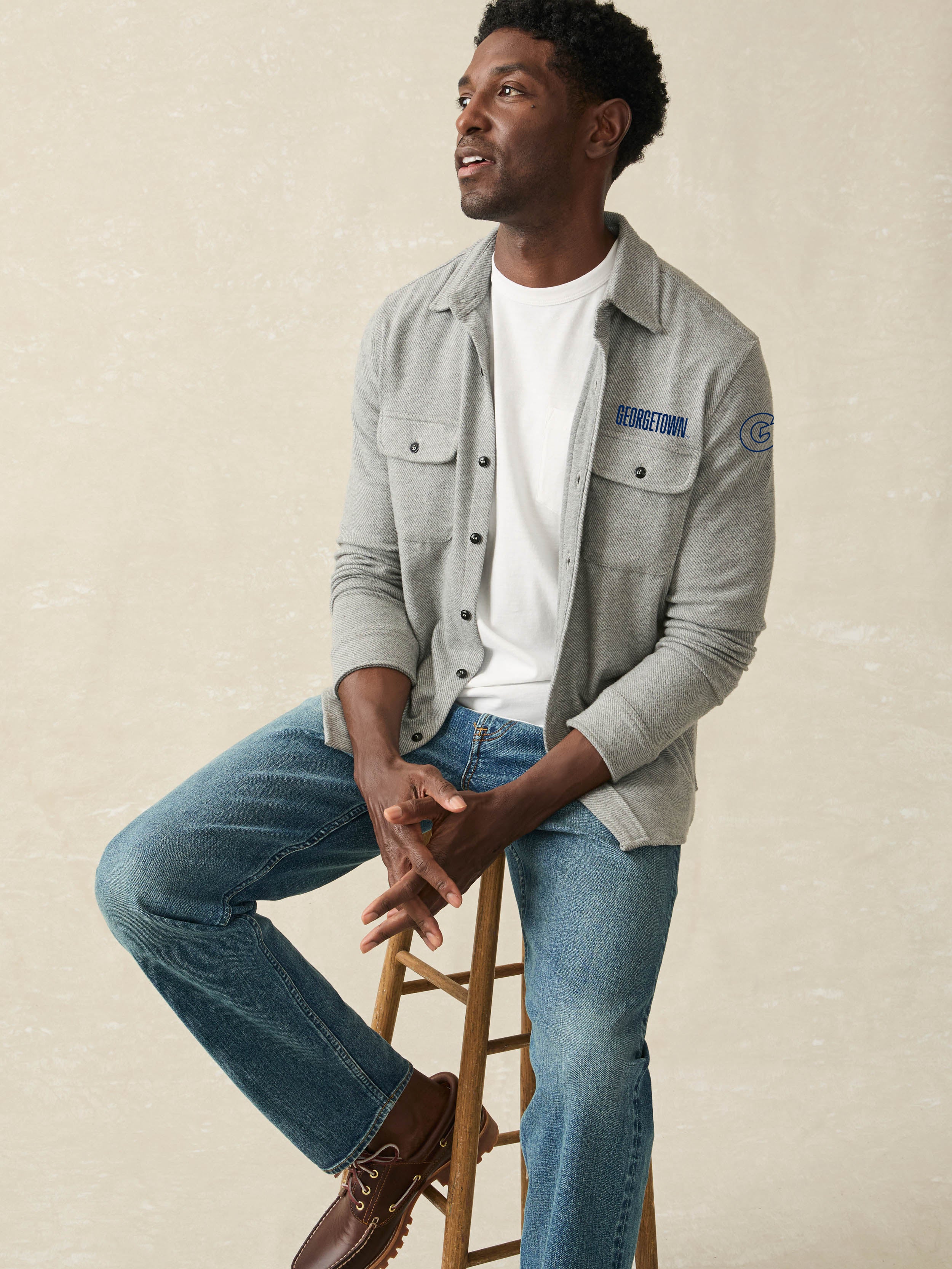Man wearing a grey long sleeve button up shirt with Georgetown graphic on the left chest and left arm against a beige background