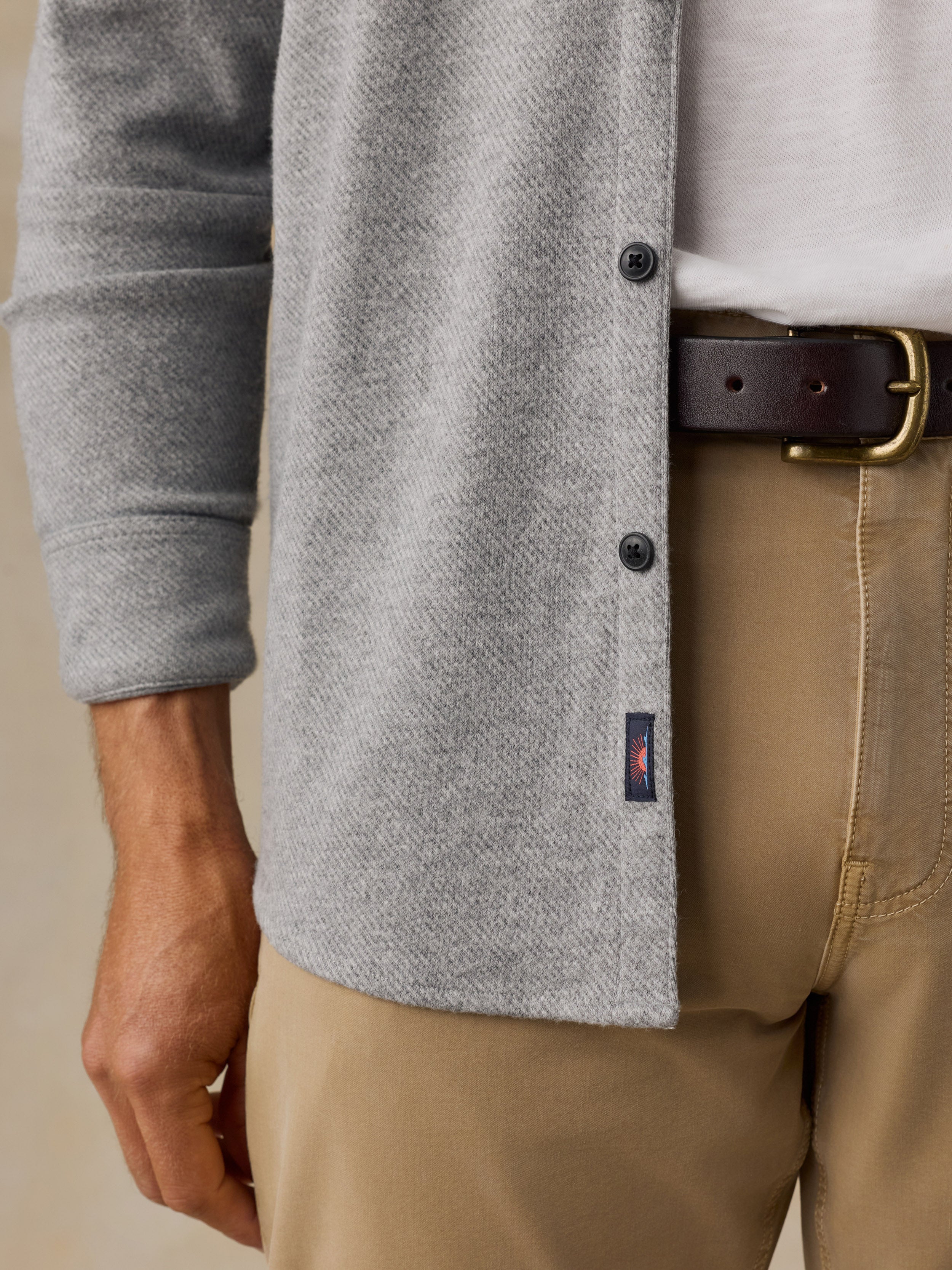 Close-up of a man wearing a light gray button-front cardigan over a white shirt, tan pants, and a dark brown belt with a gold buckle. A small navy Faherty brand tab near the hem.