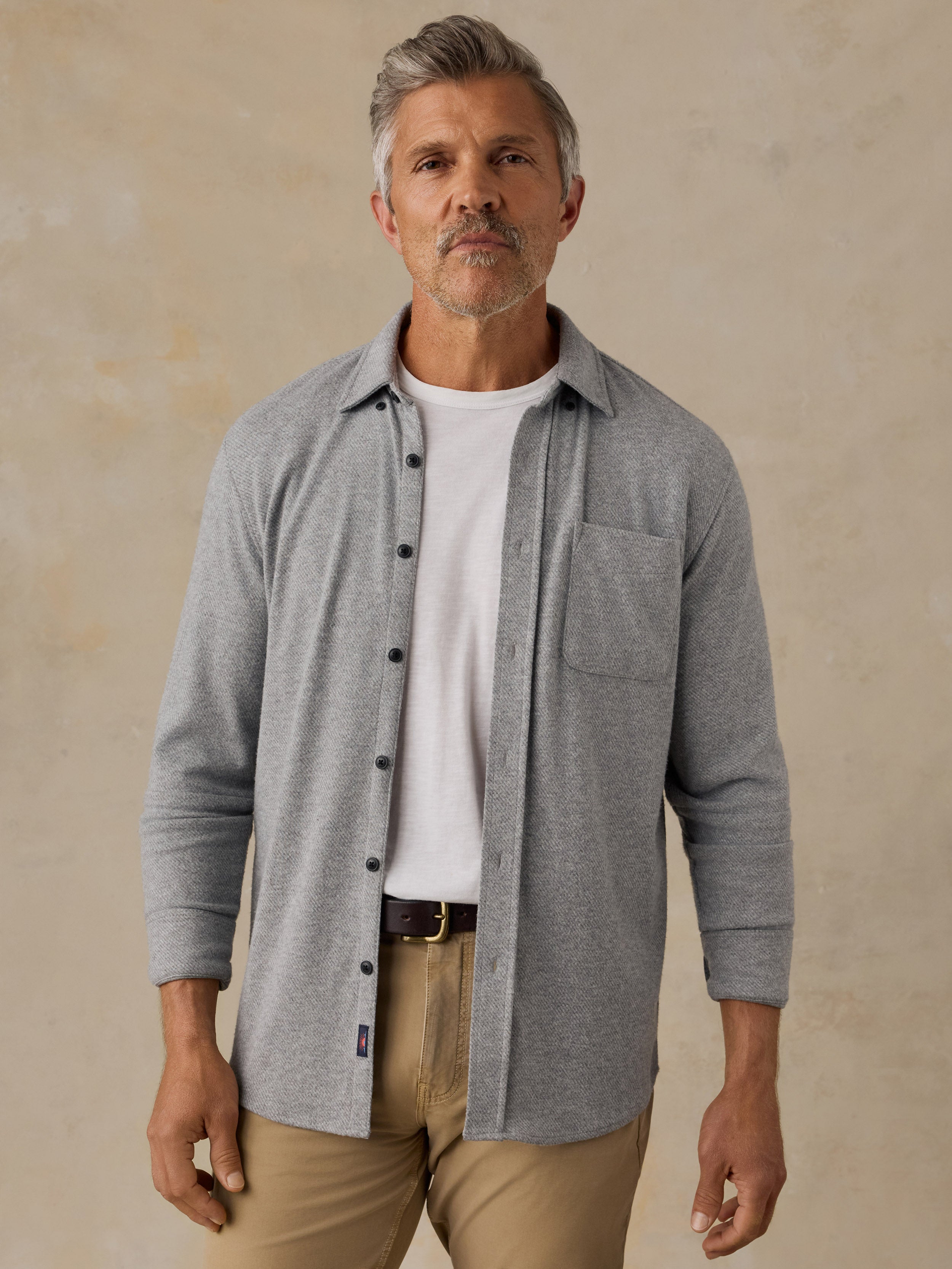A man wearing a light gray long-sleeve button-up shirt over a white t-shirt and khaki pants, standing against a textured beige background.