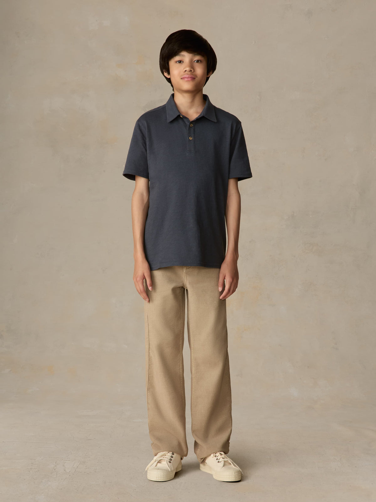 Child wearing a blue polo shirt and beige pants against a beige background, image 1 of 4