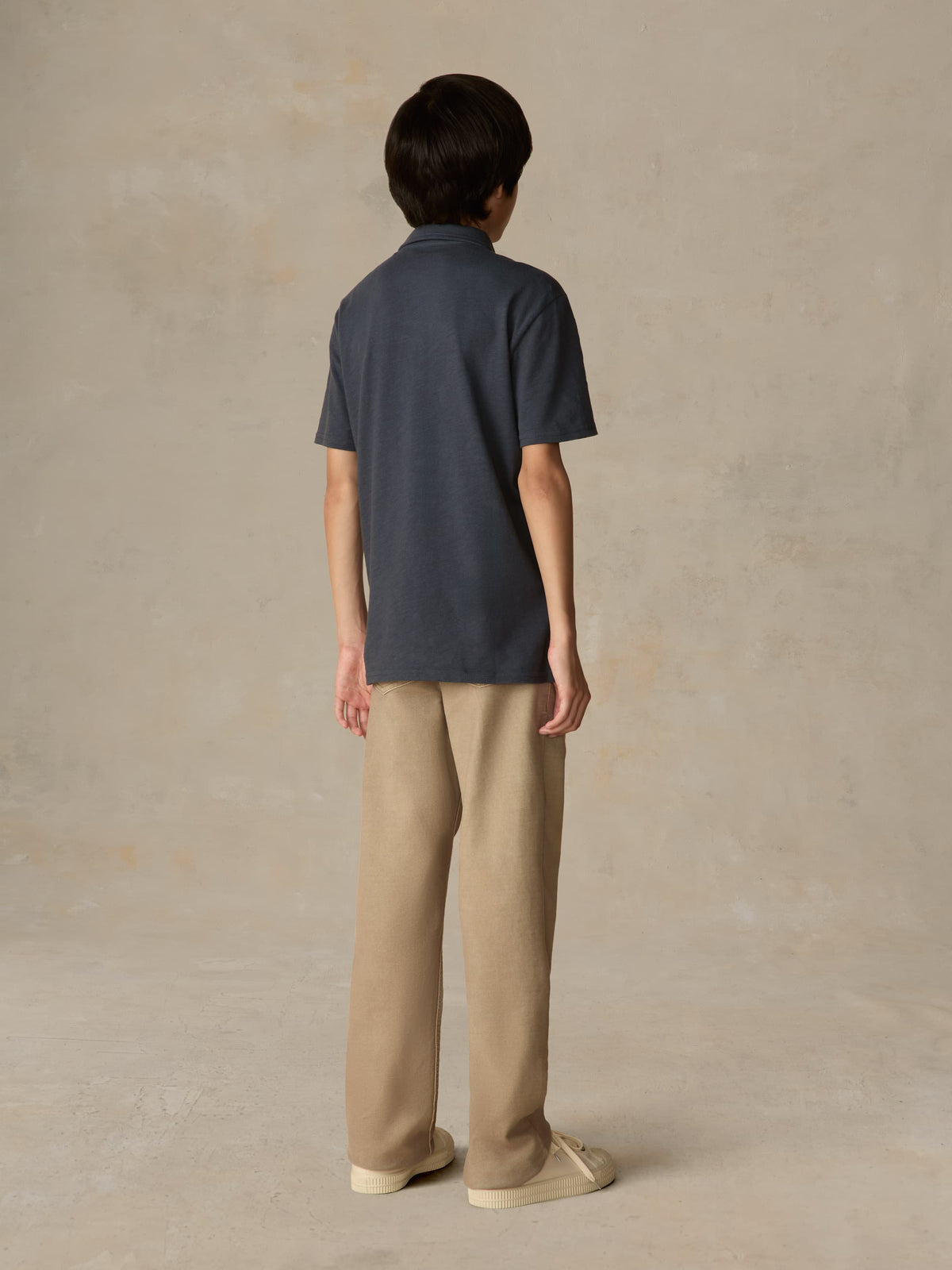 Child wearing a blue polo shirt and beige pants against a beige background, image 3 of 4