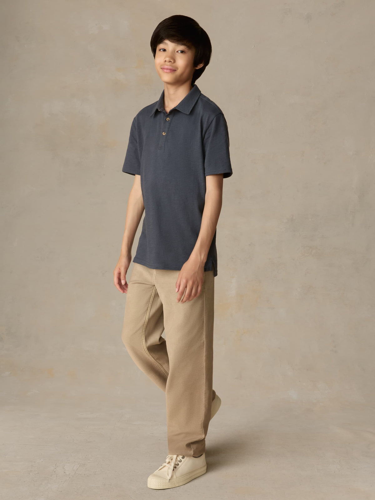 Child wearing a blue polo shirt and beige pants against a beige background, image 2 of 4