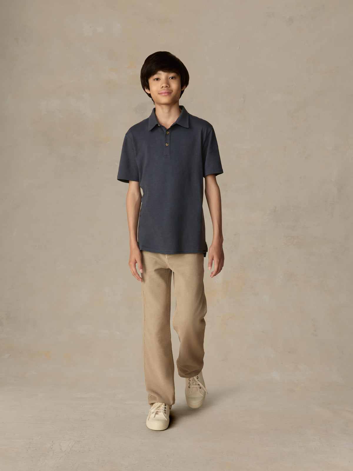 Child wearing a blue polo shirt and beige pants against a beige background, image 4 of 4