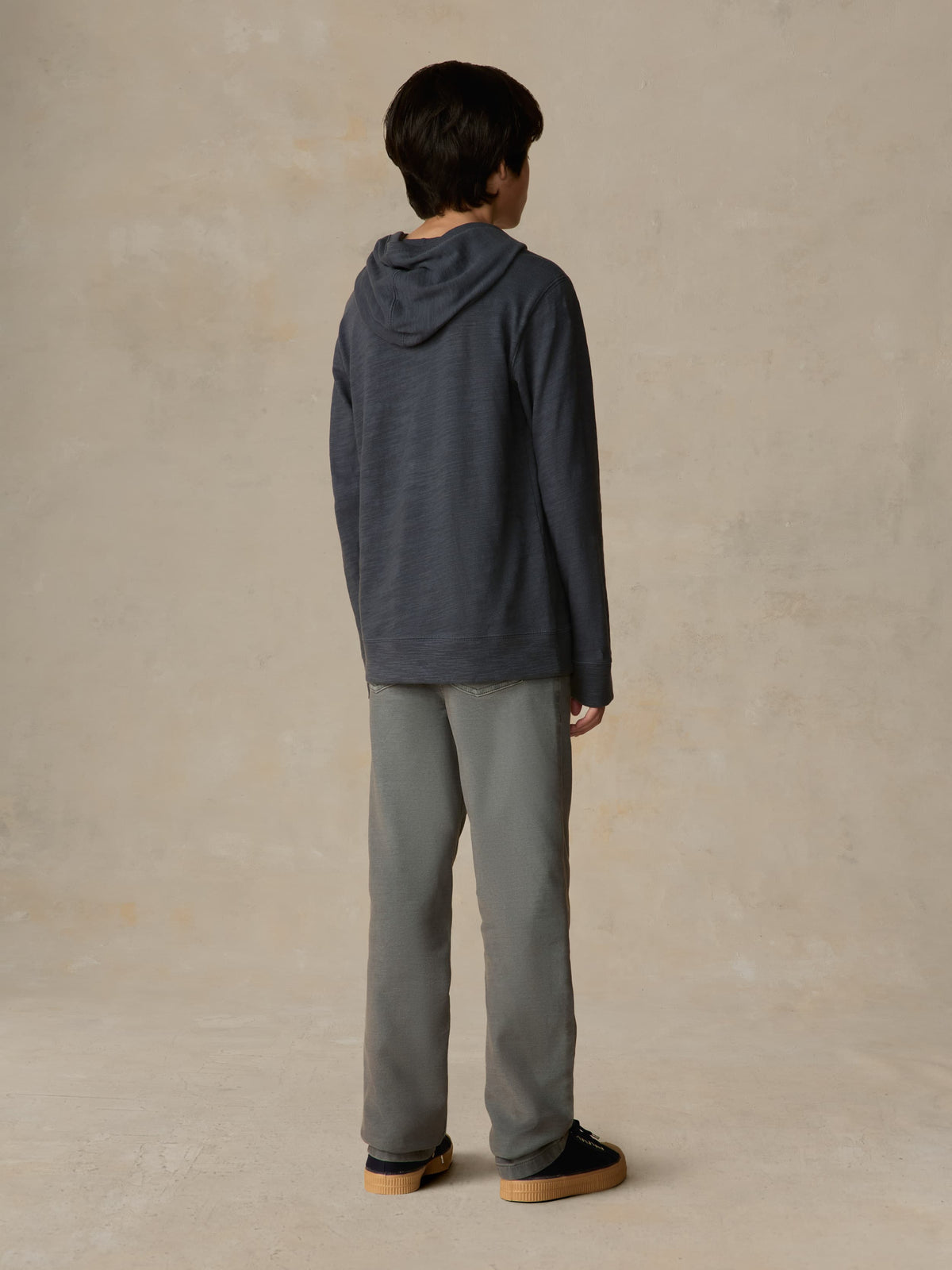 Child wearing a navy hoodie and grey pants against a beige background, image 3 of 5