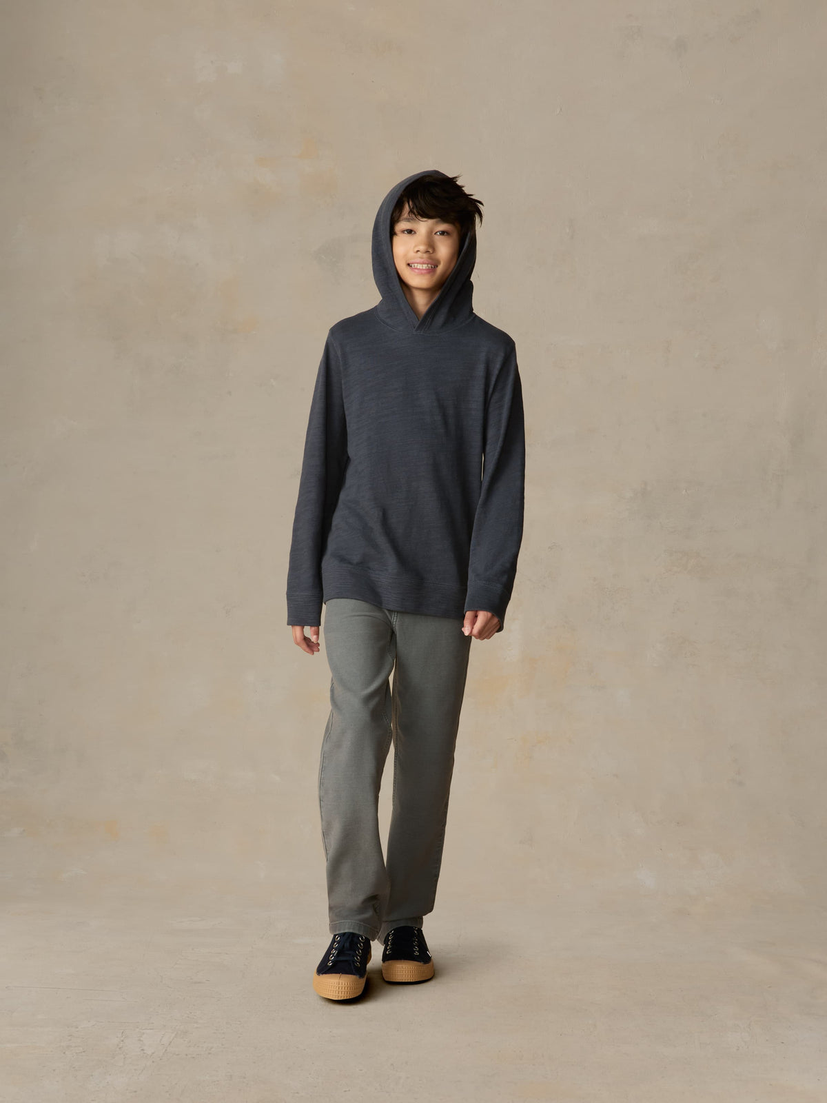 Child wearing a navy hoodie and grey pants against a beige background, image 5 of 5