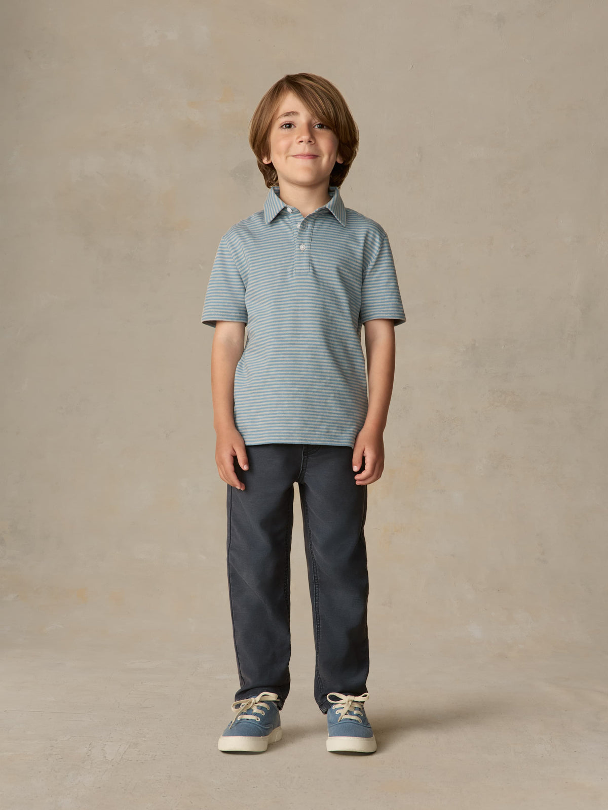 Child wearing a blue striped polo and navy pants against a beige background, image 4 of 5
