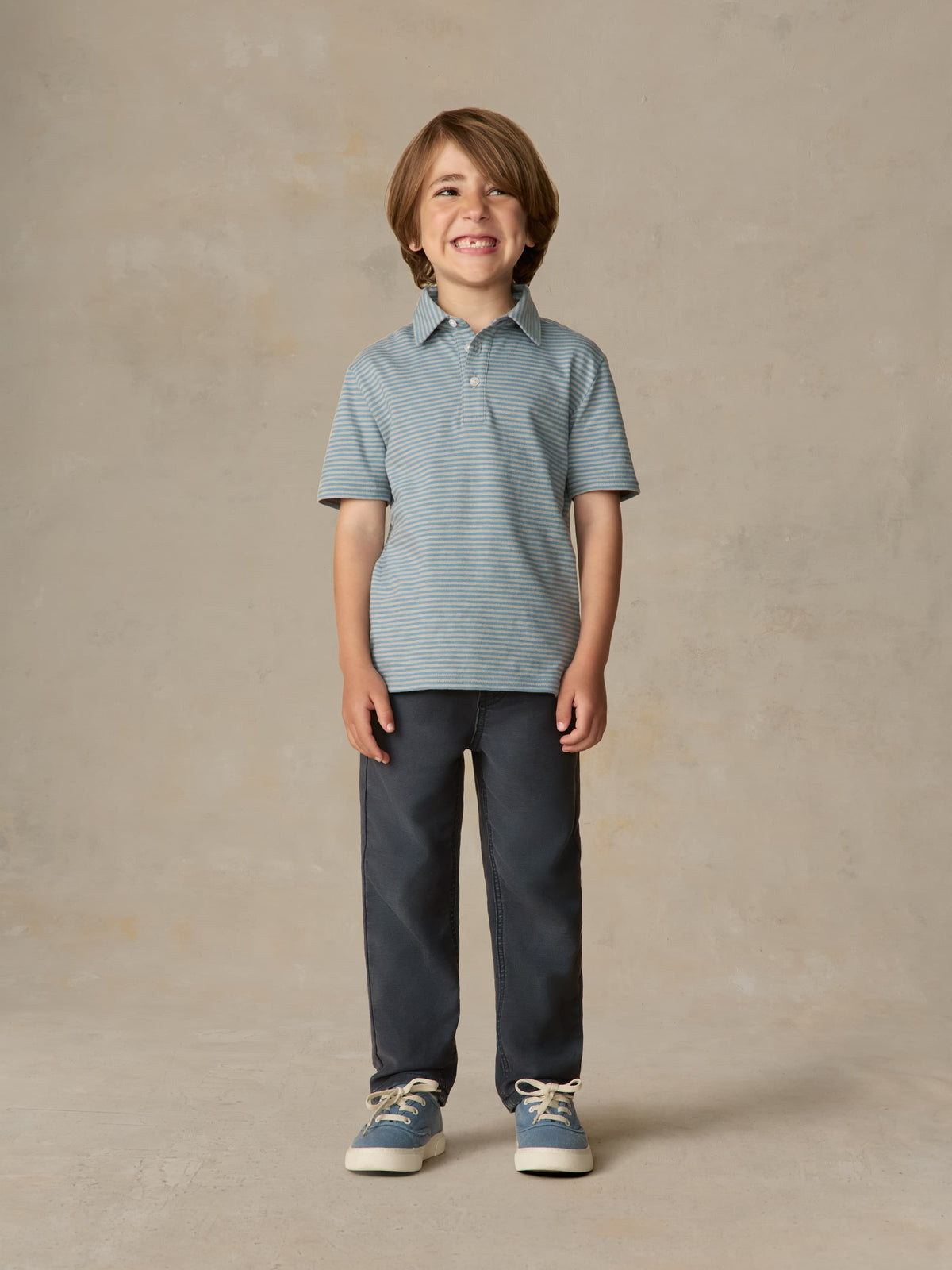 Child wearing a blue striped polo and navy pants against a beige background, image 2 of 5