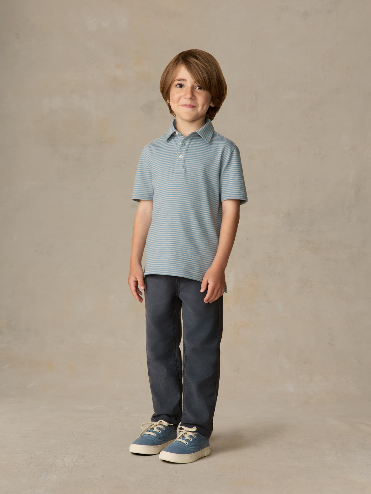Child wearing a blue striped polo and navy pants against a beige background, image 1 of 5