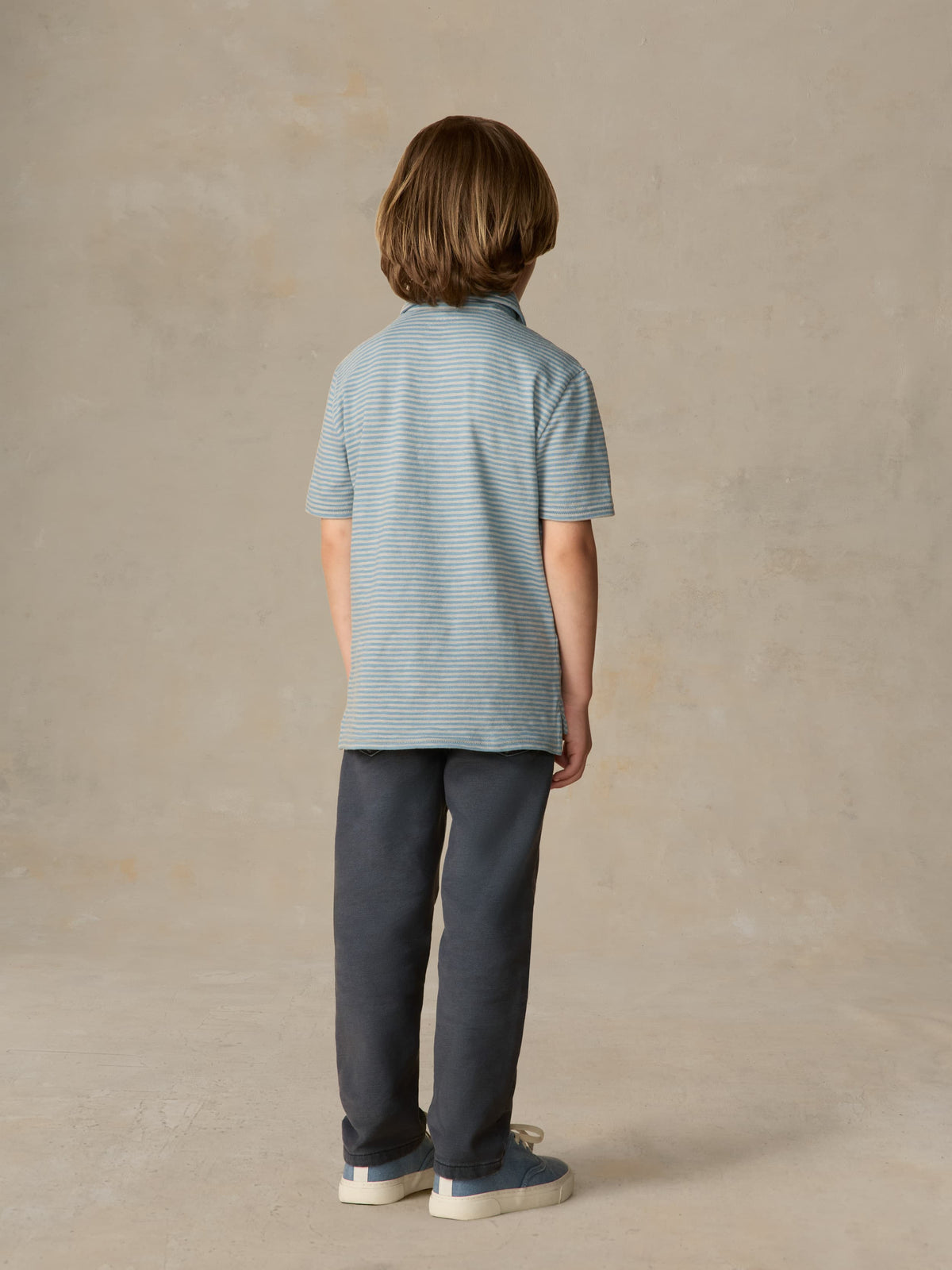 Child wearing a blue striped polo and navy pants against a beige background, image 3 of 5