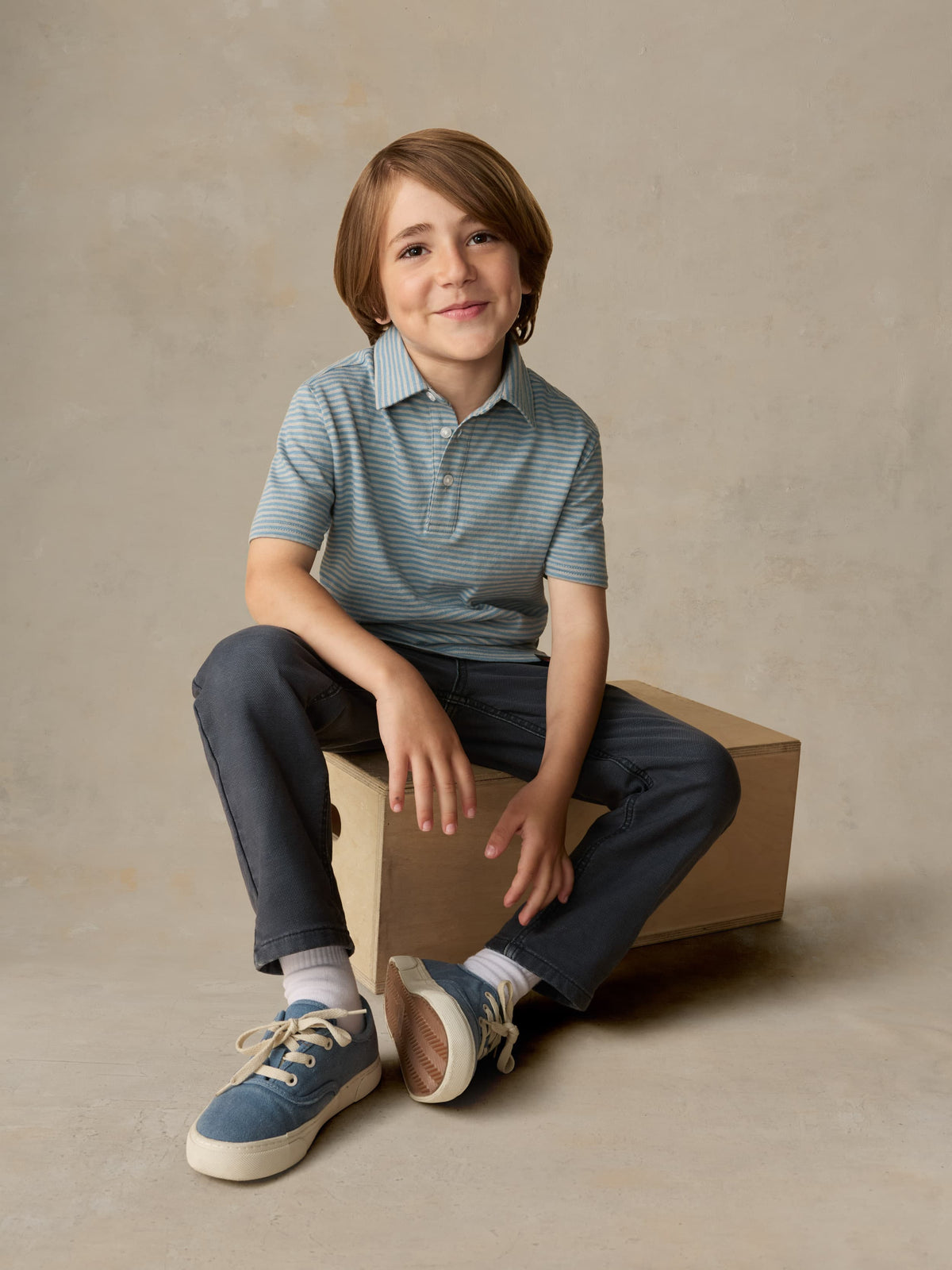 Child sitting on a wooden box wearing a blue striped polo and navy pants against a beige background, image 5 of 5