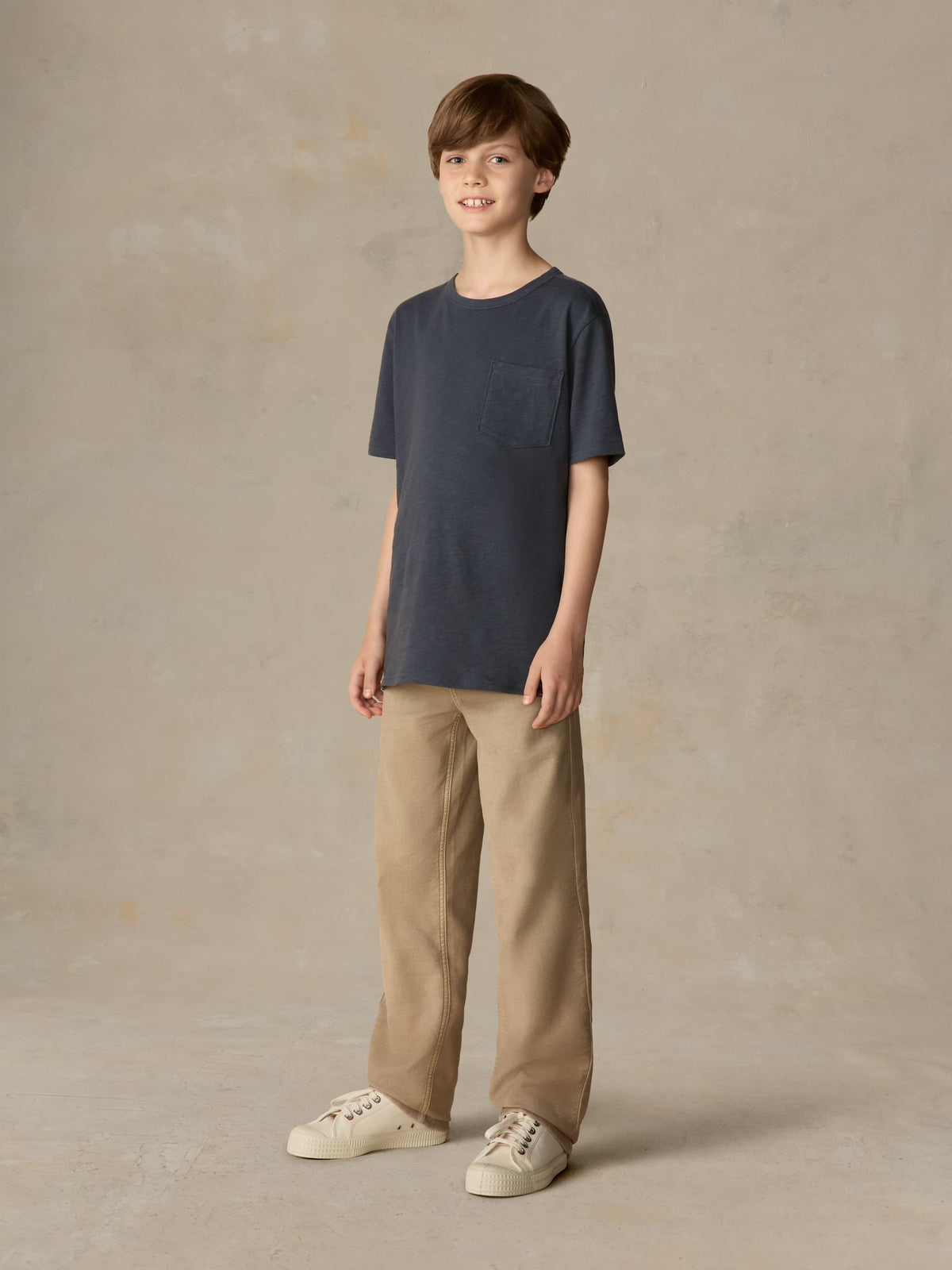 Child wearing a blue t-shirt and beige pants against a beige background, image 4 of 7