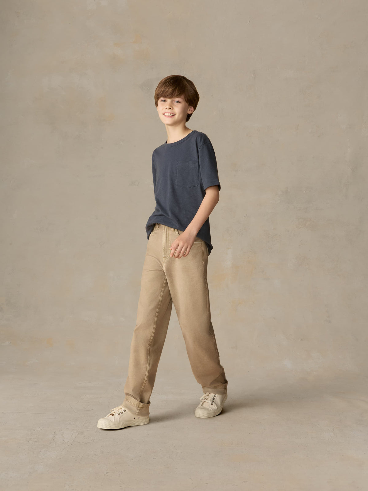Child wearing a blue t-shirt and beige pants against a beige background, image 2 of 7