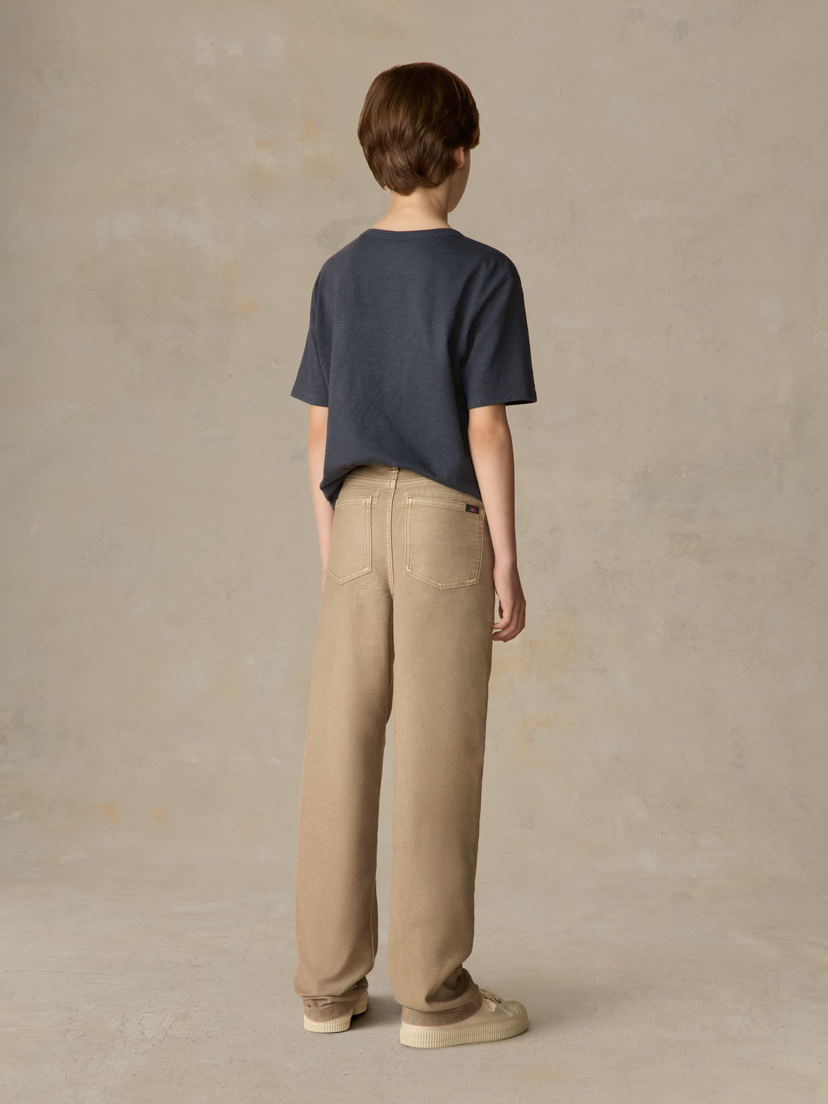 Child wearing a blue t-shirt and beige pants against a beige background, image 3 of 7