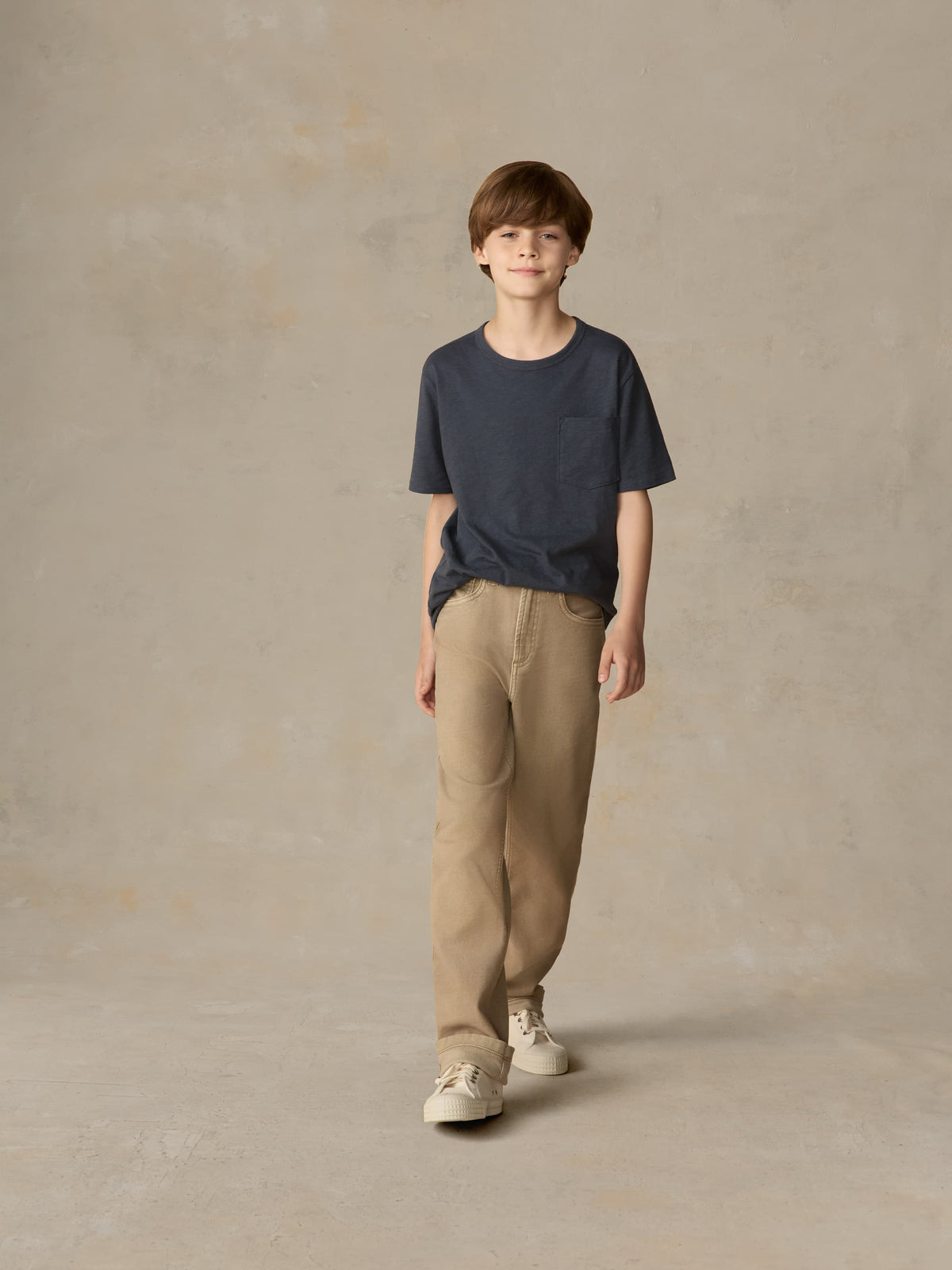 Child wearing a blue t-shirt and beige pants against a beige background, image 1 of 7