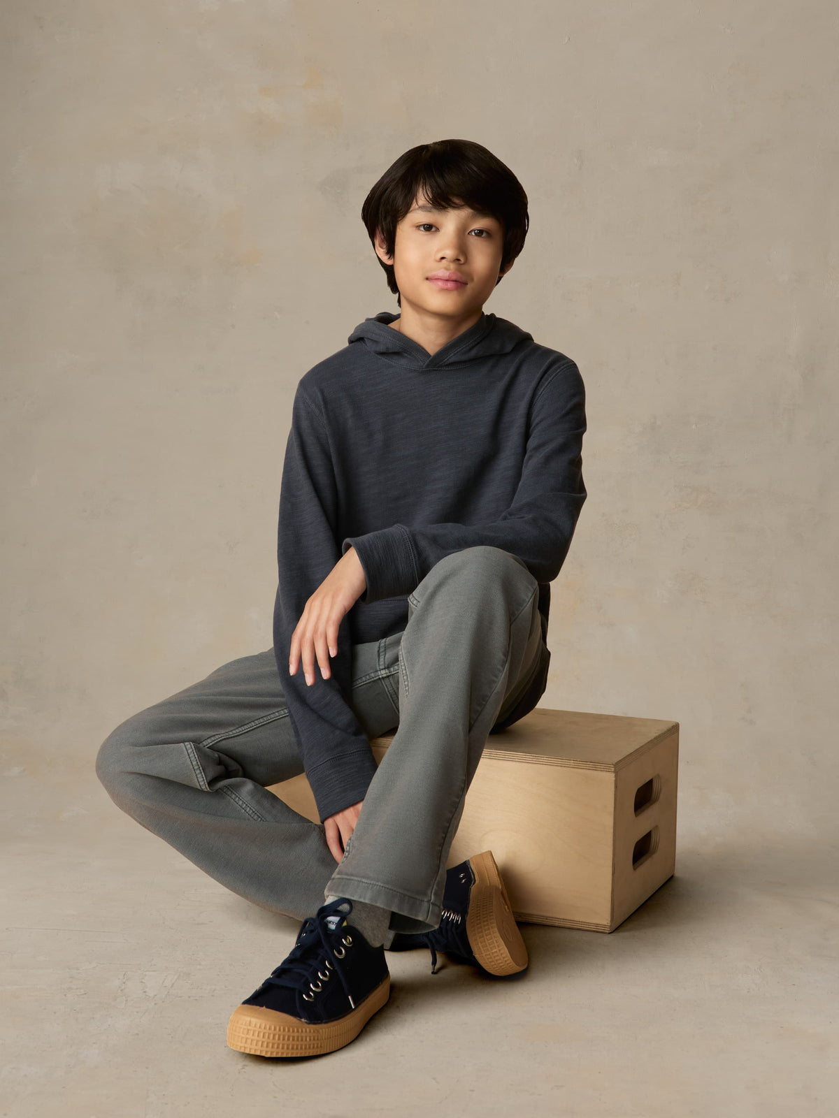 Child sitting on a wooden box wearing a blue hoodie with grey pants against a beige background, image 1 of 6