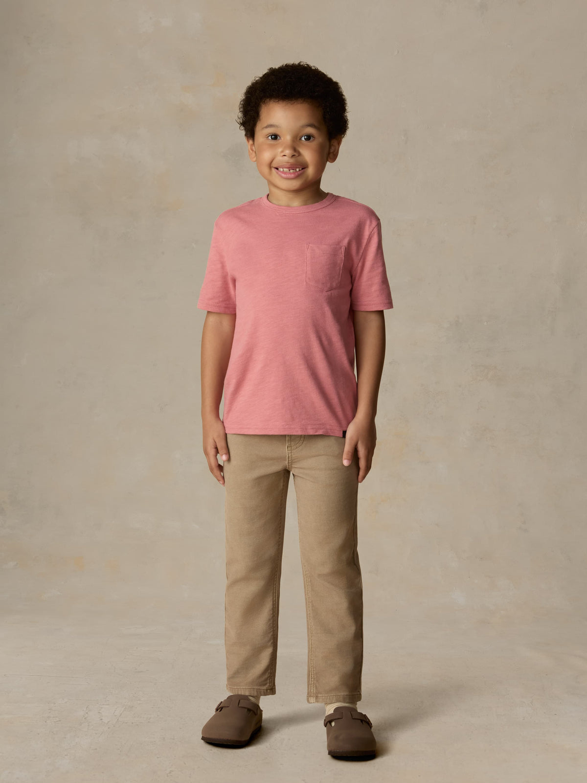 Child wearing a red t-shirt and beige pants against a beige background, image 1 of 6