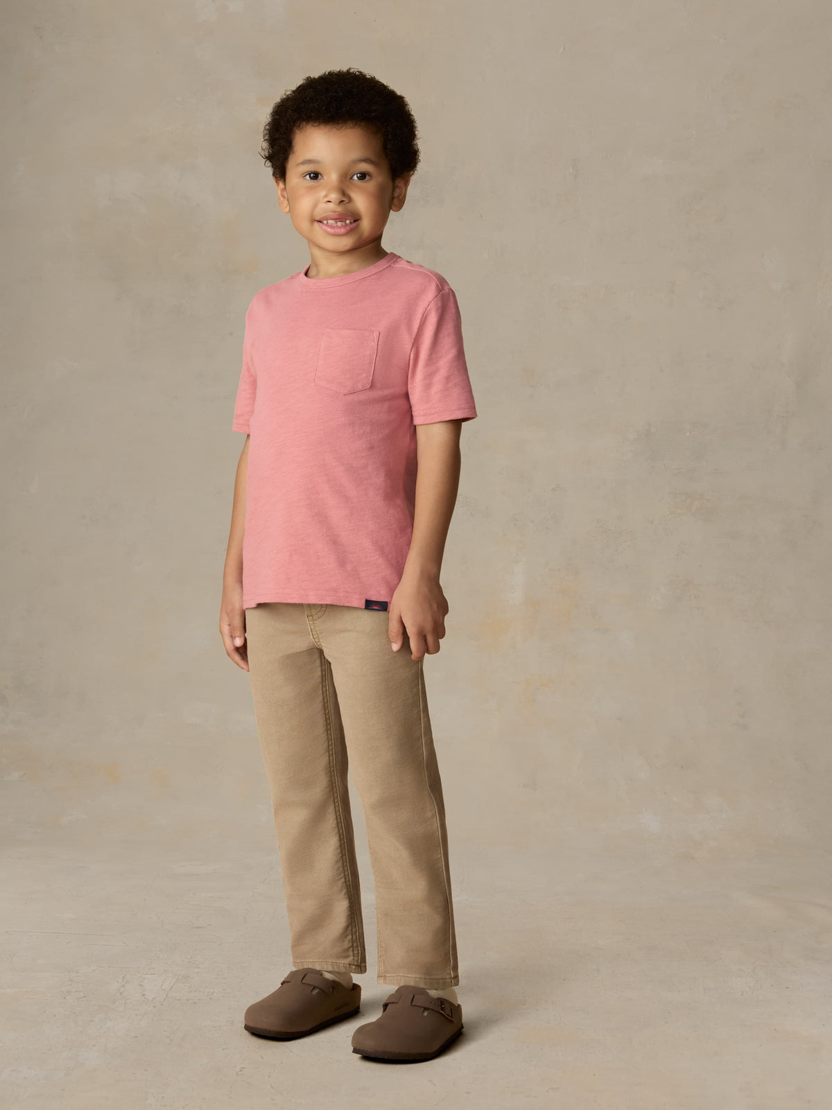 Child wearing a red t-shirt and beige pants against a beige background, image 2 of 6
