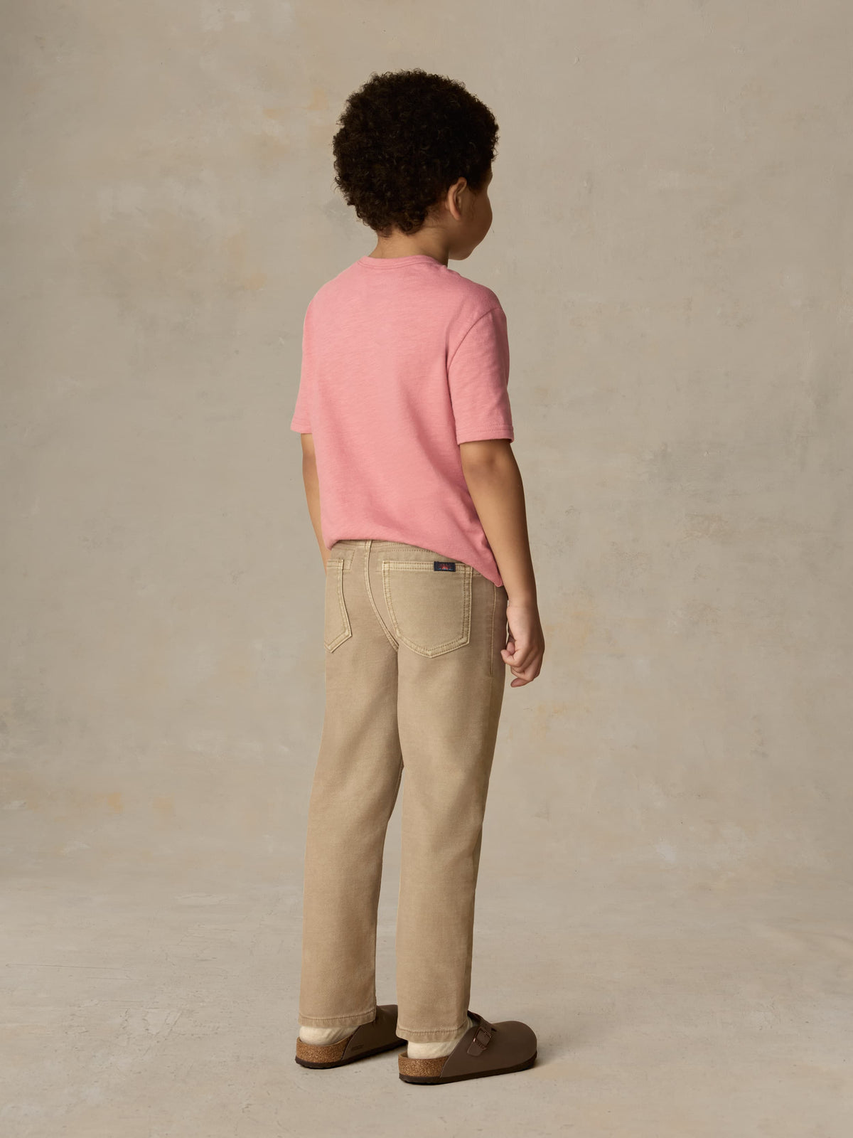 Child wearing a red t-shirt and beige pants against a beige background, image 3 of 6