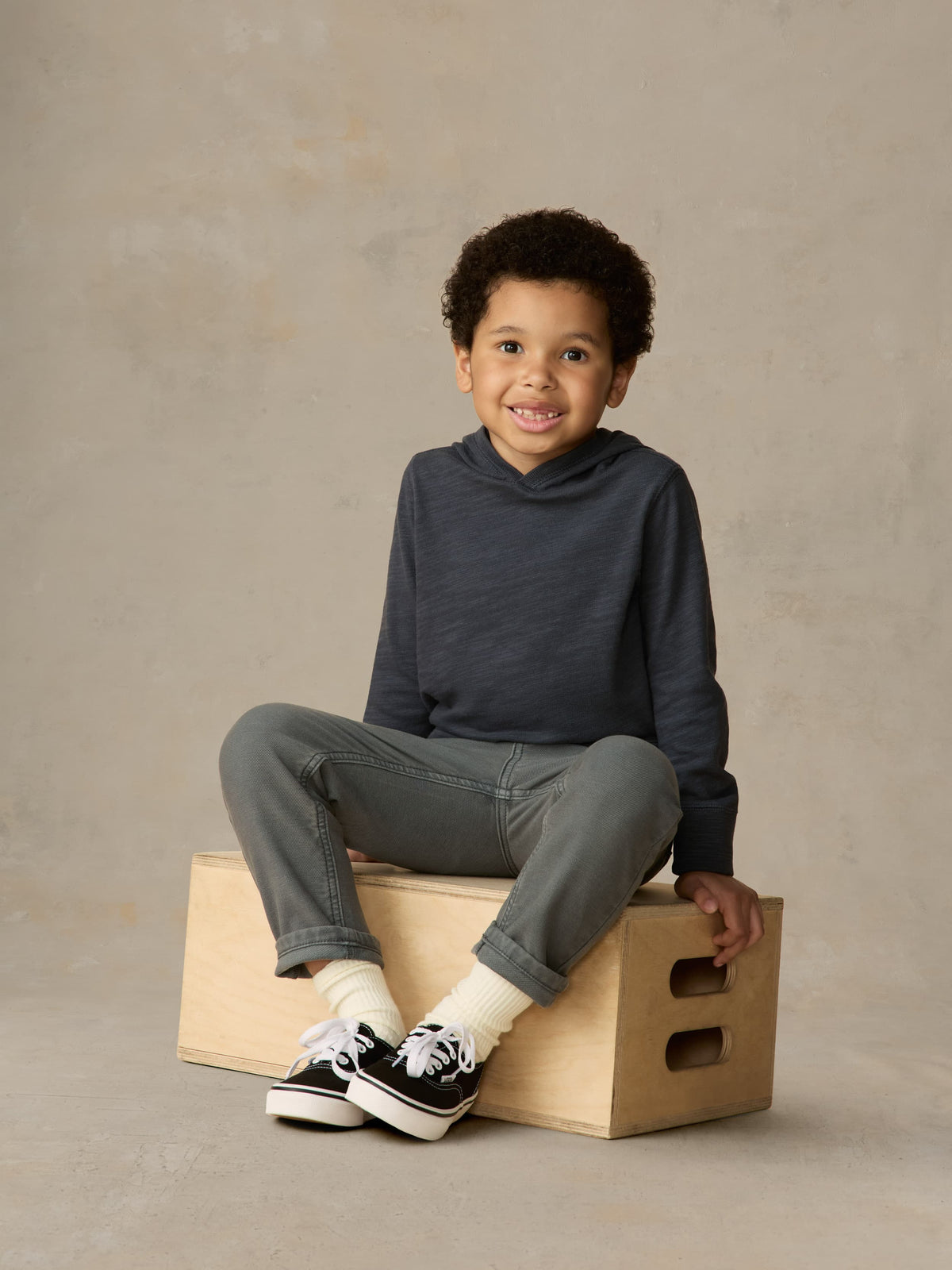 Child sitting on a wooden box wearing blue hoodie and grey pants against a beige background, image 3 of 6