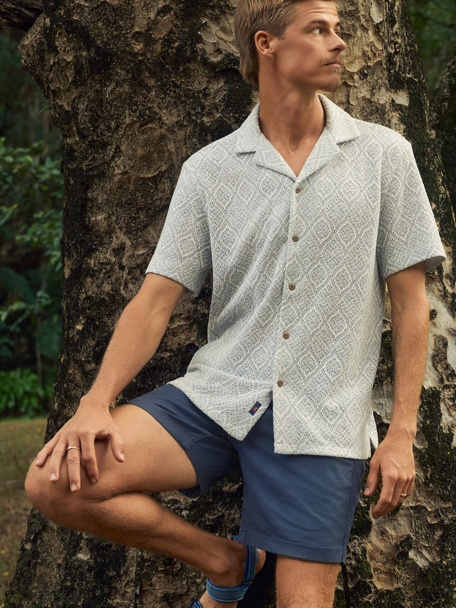 Man wearing a patterned shirt and blue shorts leaning against a tree