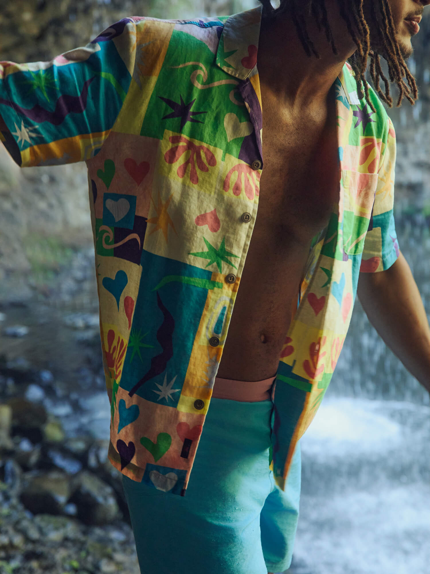 Man wearing a colorful printed shirt and swim shorts near a waterfall.