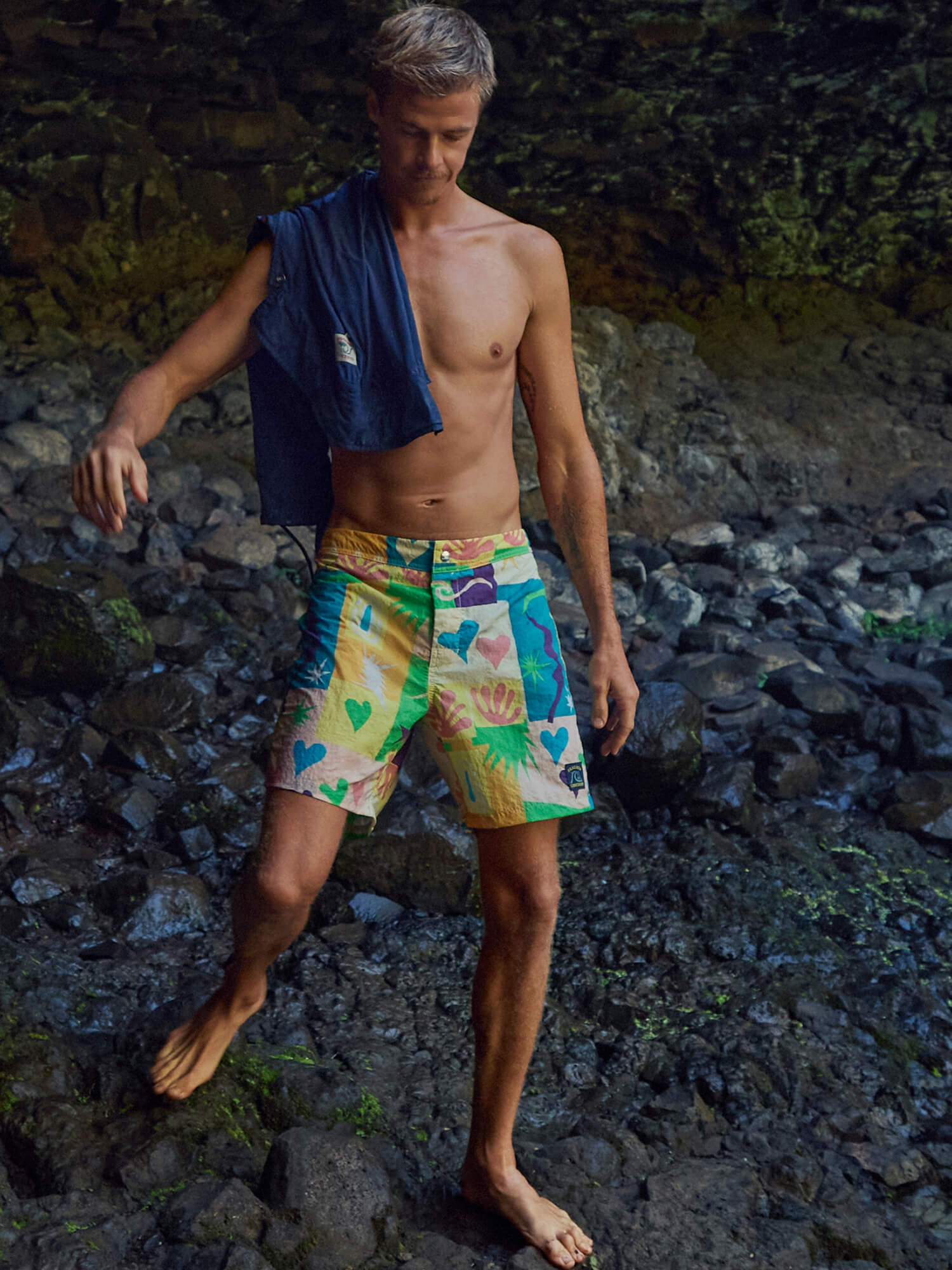 Man wearing colorful swim shorts standing on a rocky surface with a jacket draped over his shoulder.