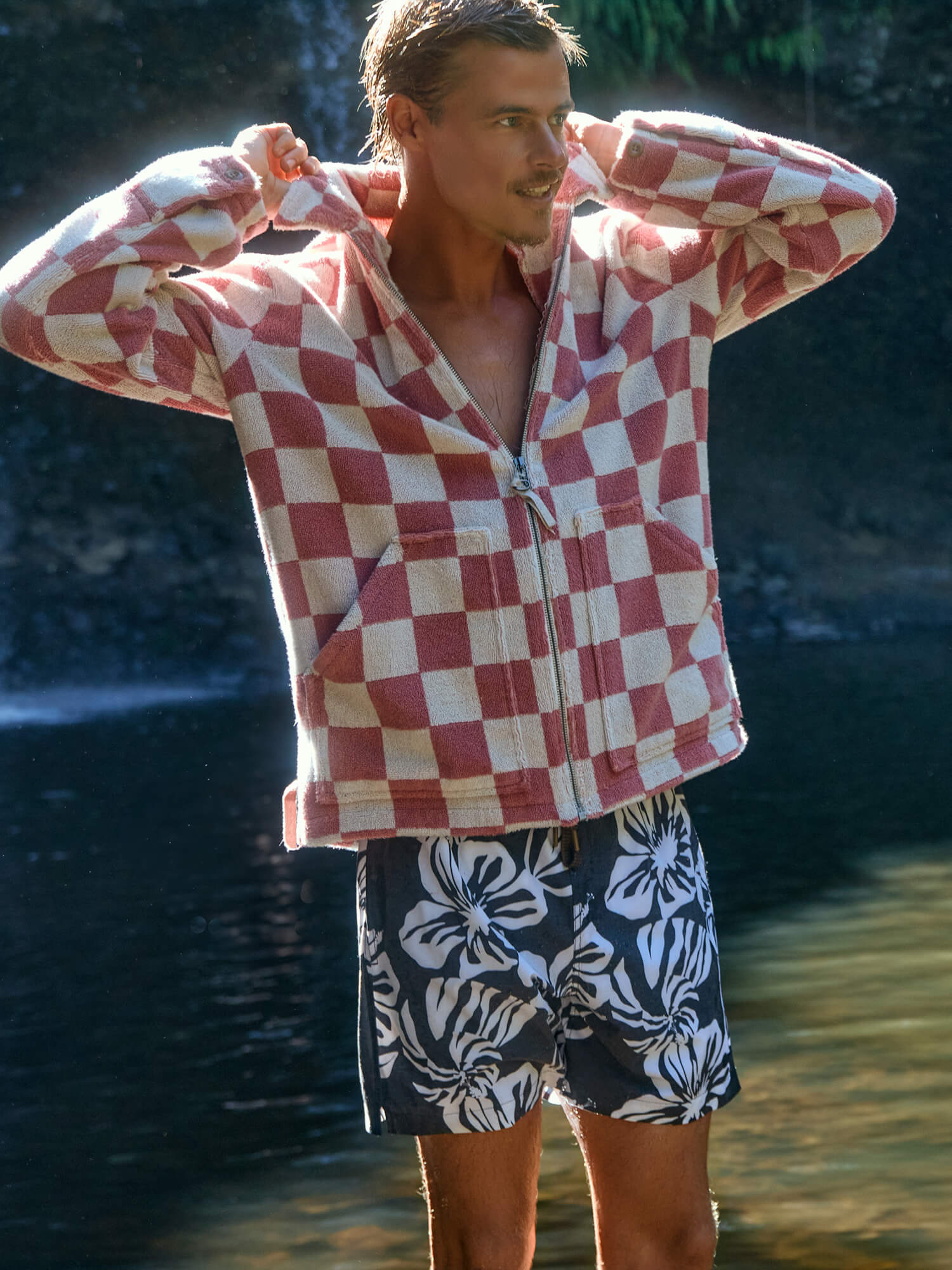 Man wearing a red and white checkered jacket and blue floral printed swim shorts by water.