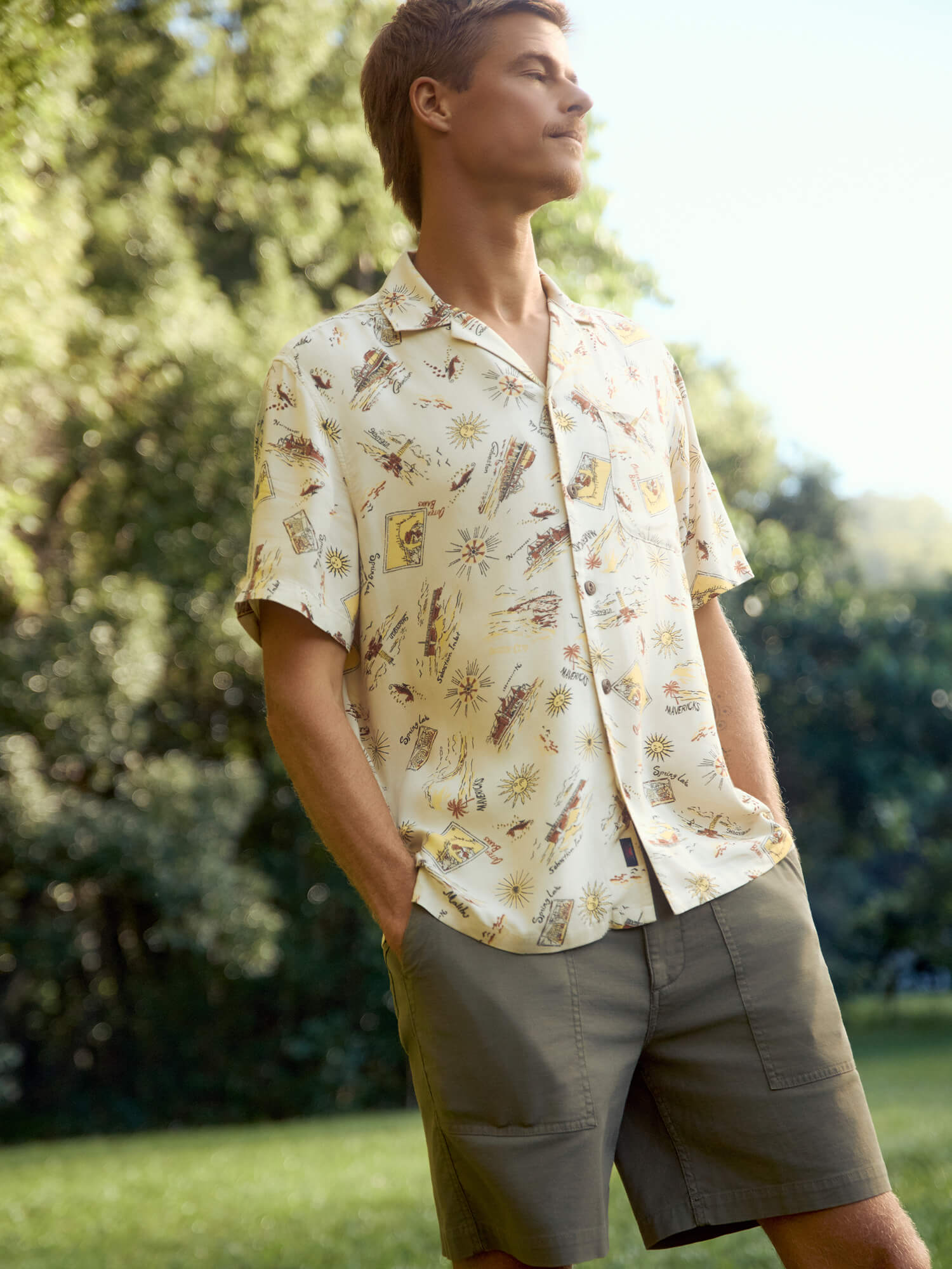 Man wearing a patterned shirt and shorts standing outdoors with greenery in the background