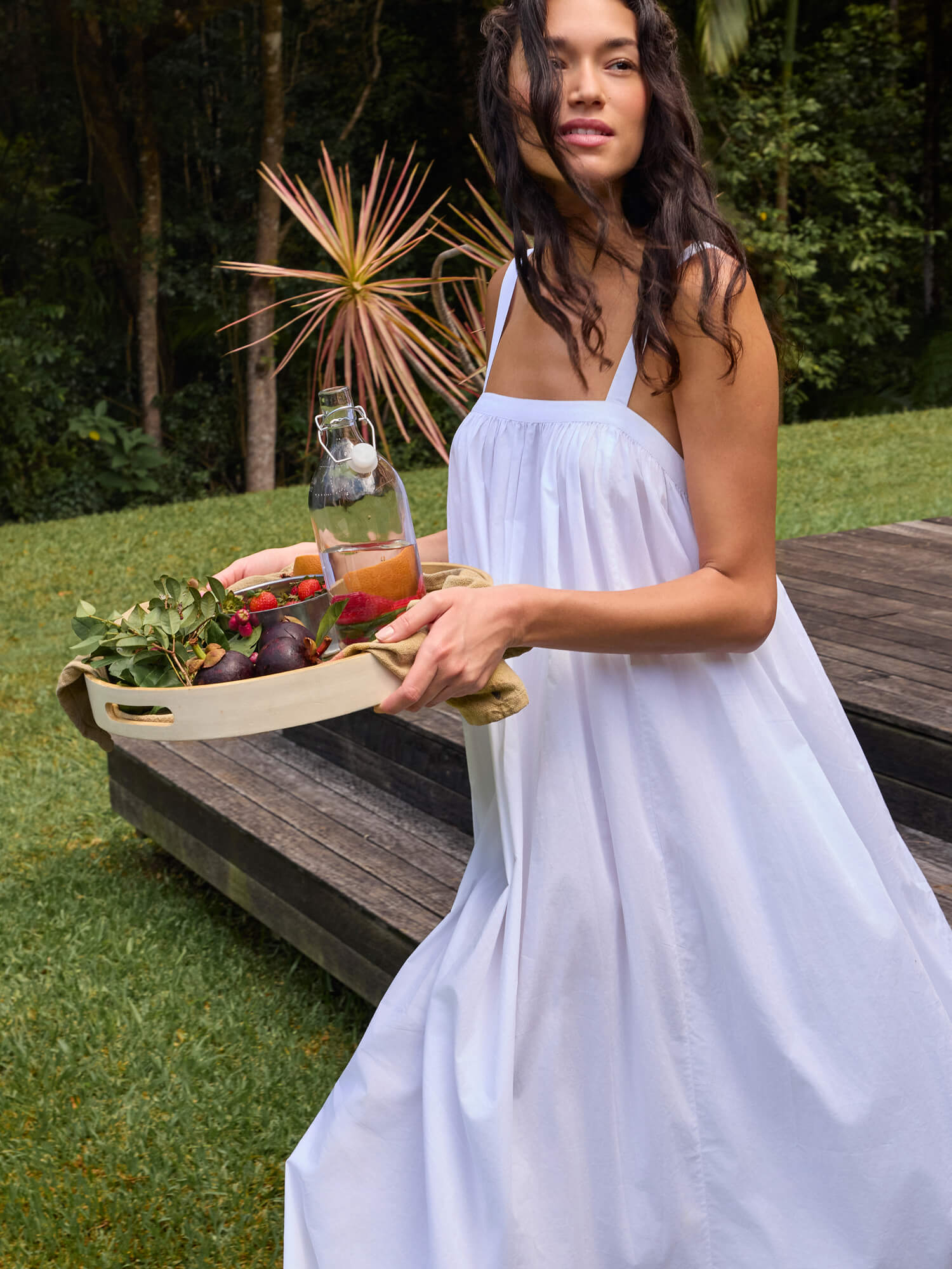 Woman in a white dress holding a tray with food and water outdoors.
