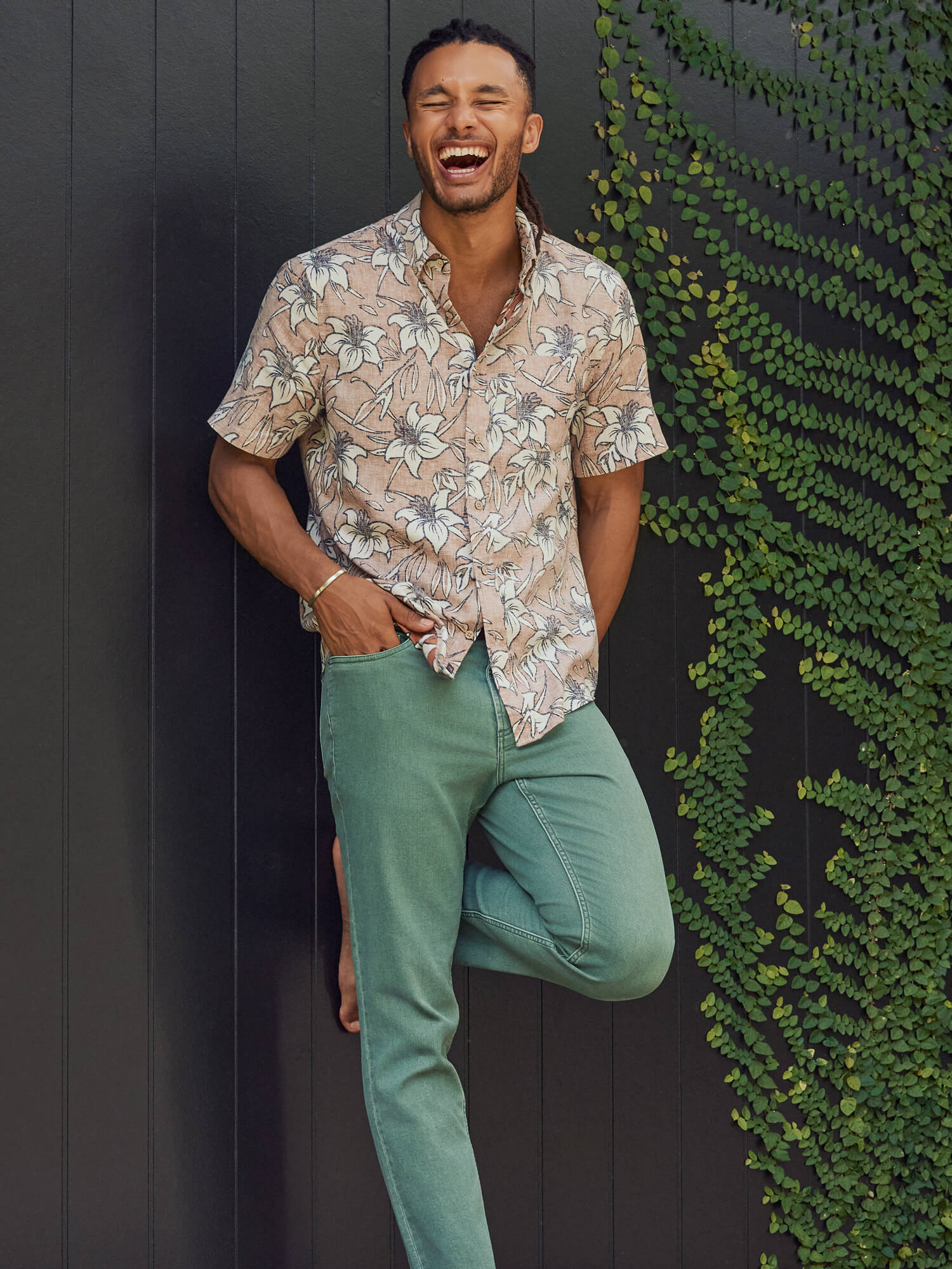 Man in a floral shirt and green pants leaning against a wall with greenery.