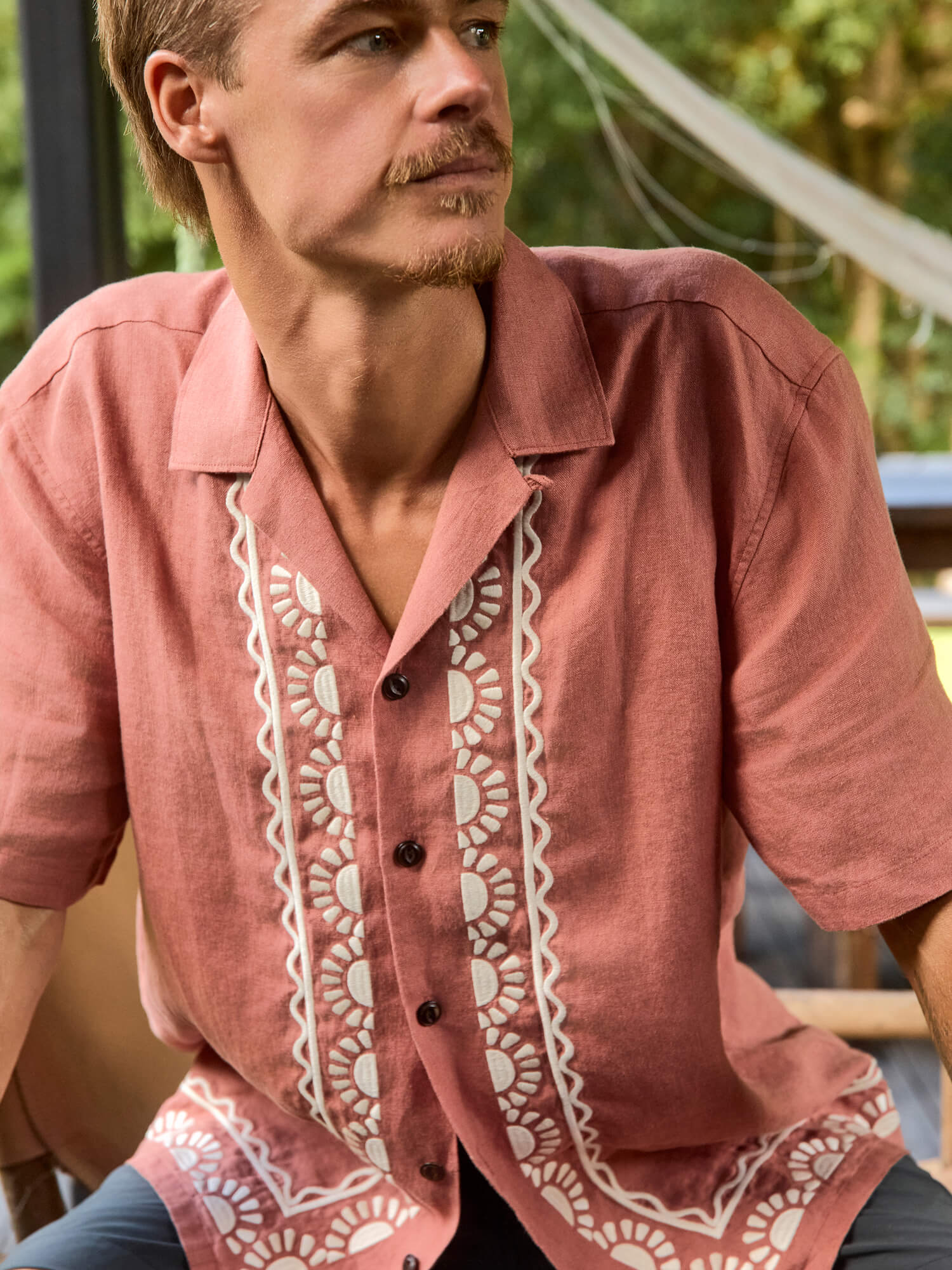 Man wearing an pale red shirt with white embroidery sitting outdoors.