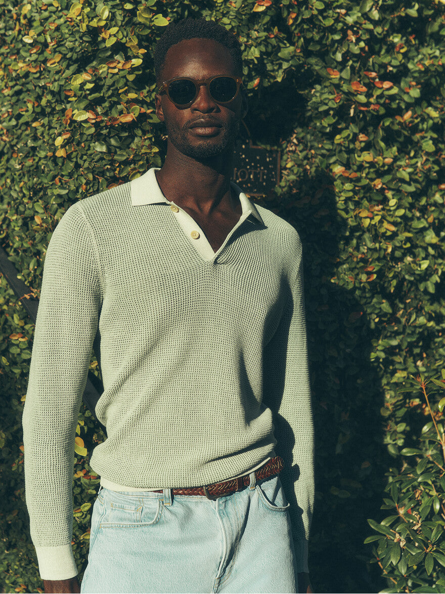 Man posing in front of a hedge wearing sunglasses, a knit sweater polo shirt, a braided belt and sunwashed indigo stretch terry pants